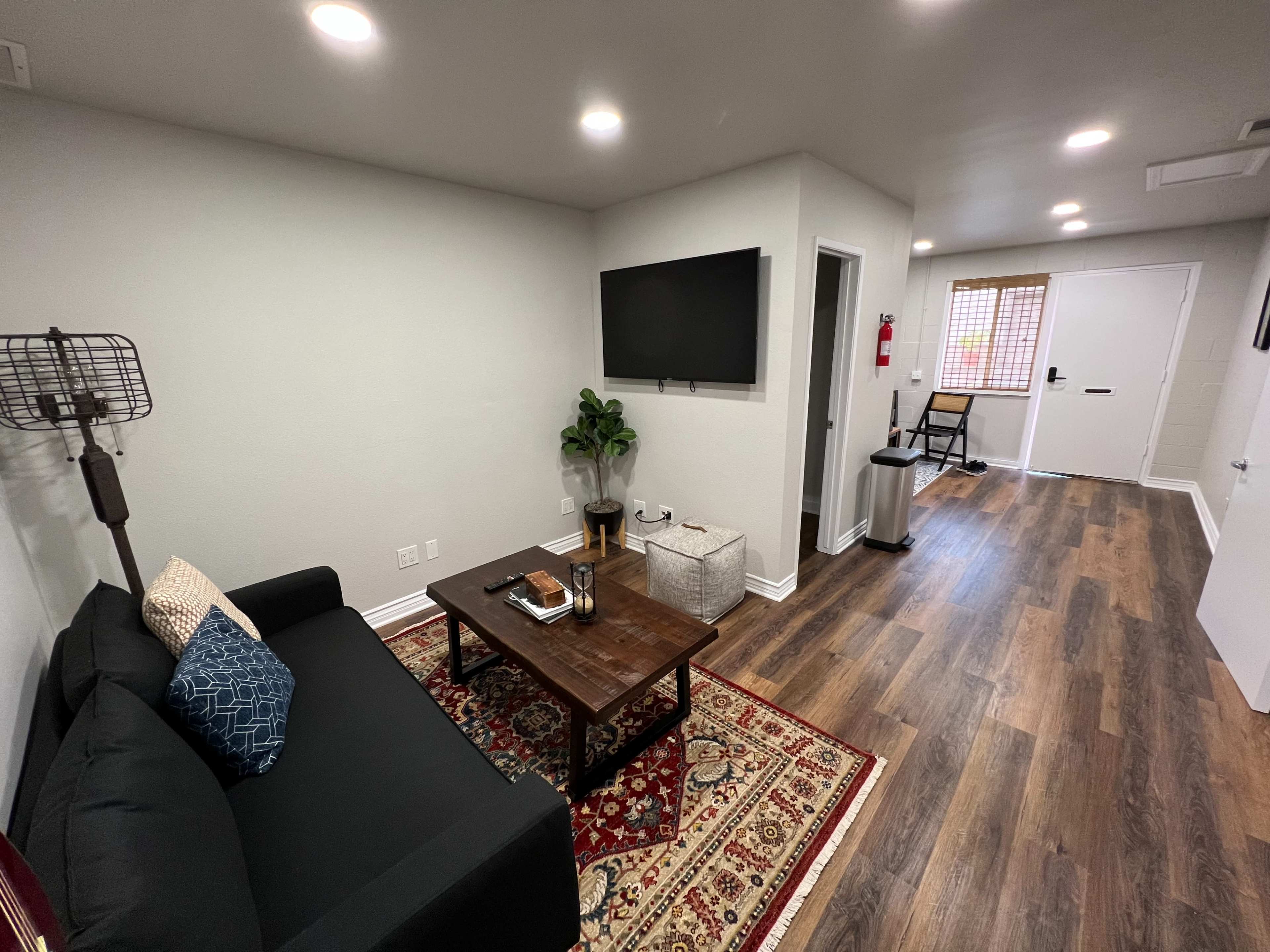 The image shows a small living area with a black couch, a wooden coffee table, and a television mounted on the wall, leading to a door and a window in the background.