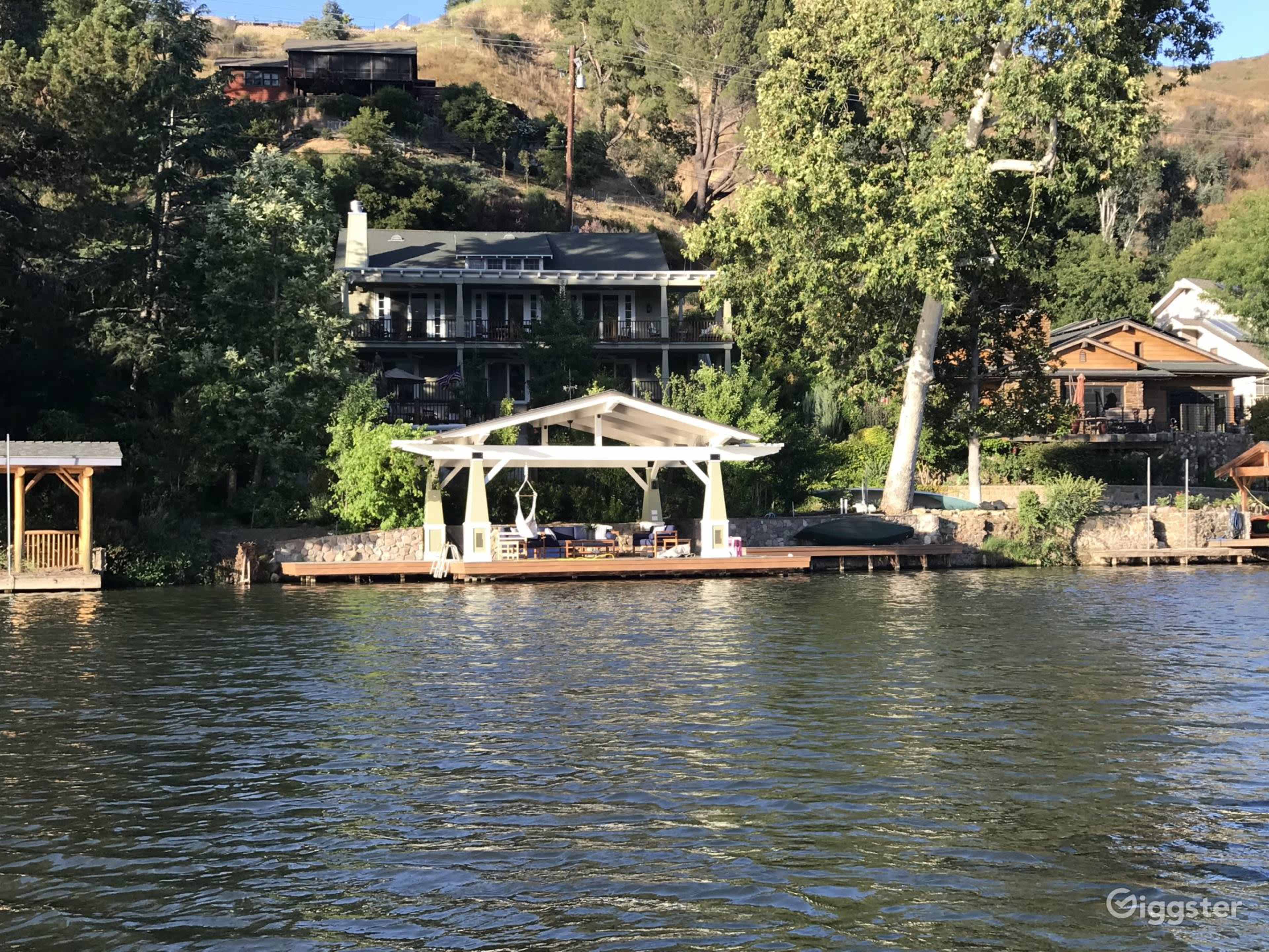 A waterfront scene with a dock featuring a covered seating area and several houses nestled on a hill behind it.