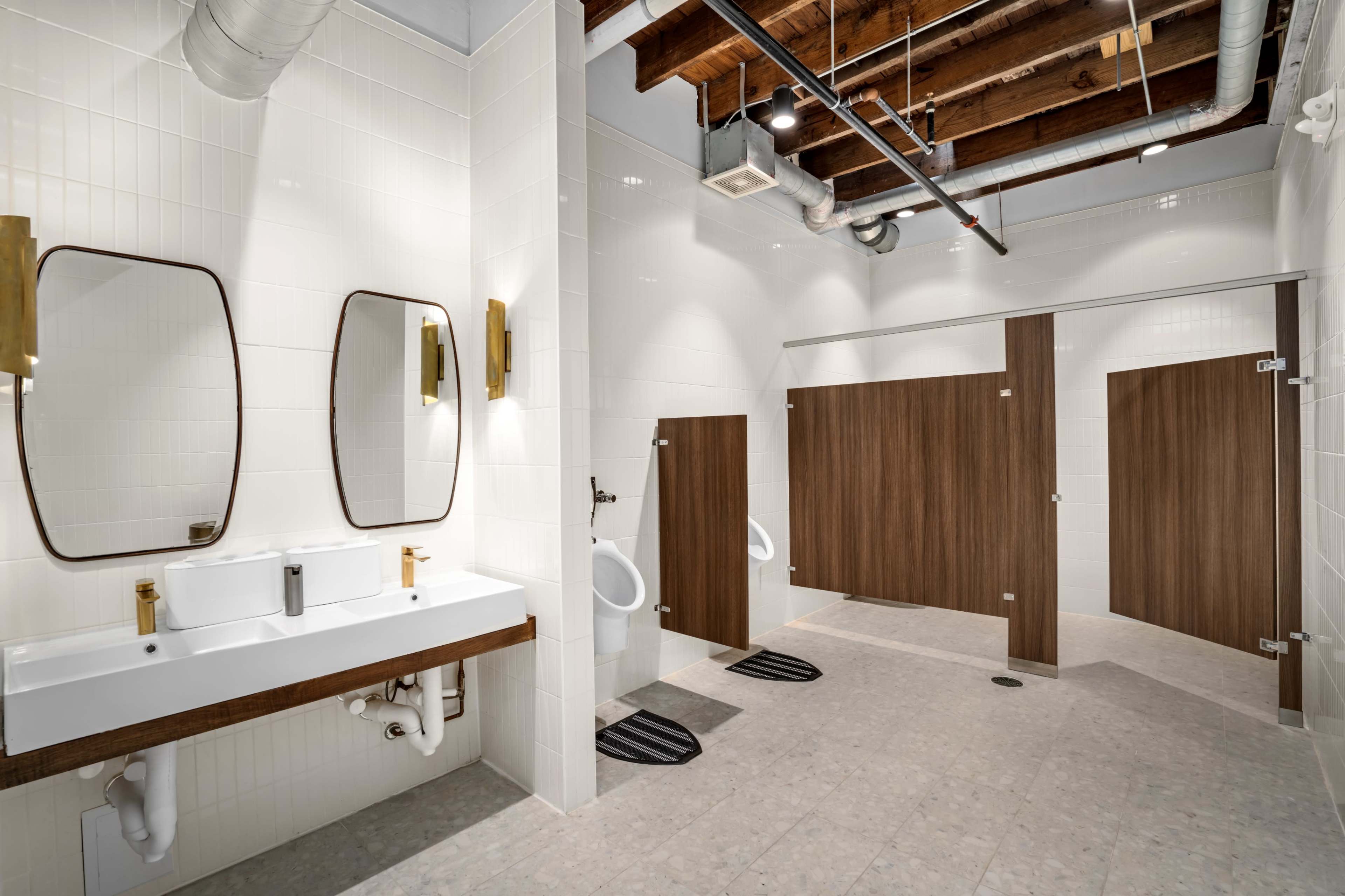 The image shows a modern restroom with two sinks, large mirrors, and separate stalls for toilets, featuring wooden doors and tiled walls.