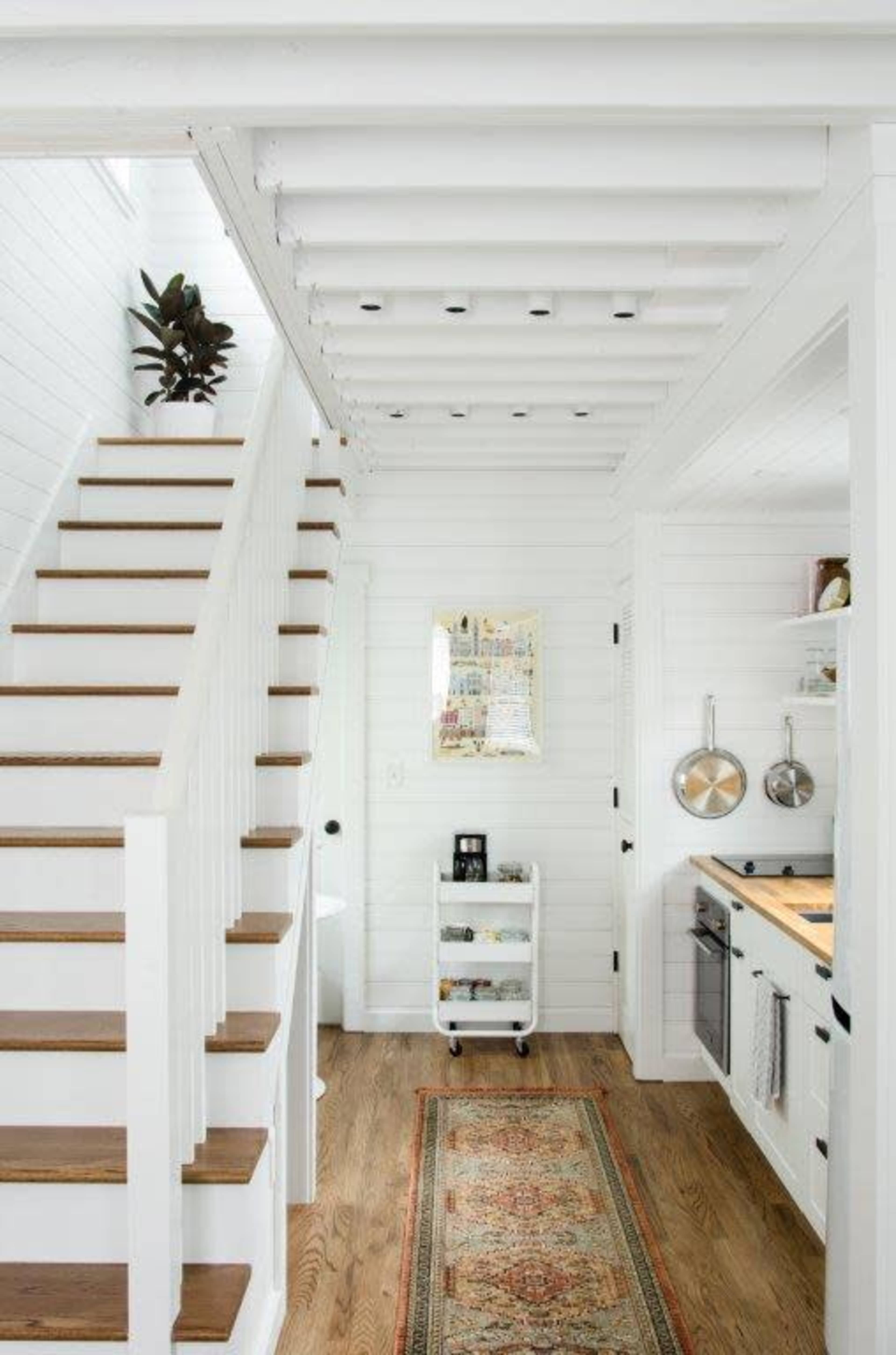 The image shows a bright, minimalist interior featuring wooden stairs, white walls, a small kitchen area, and a colorful rug on a hardwood floor.