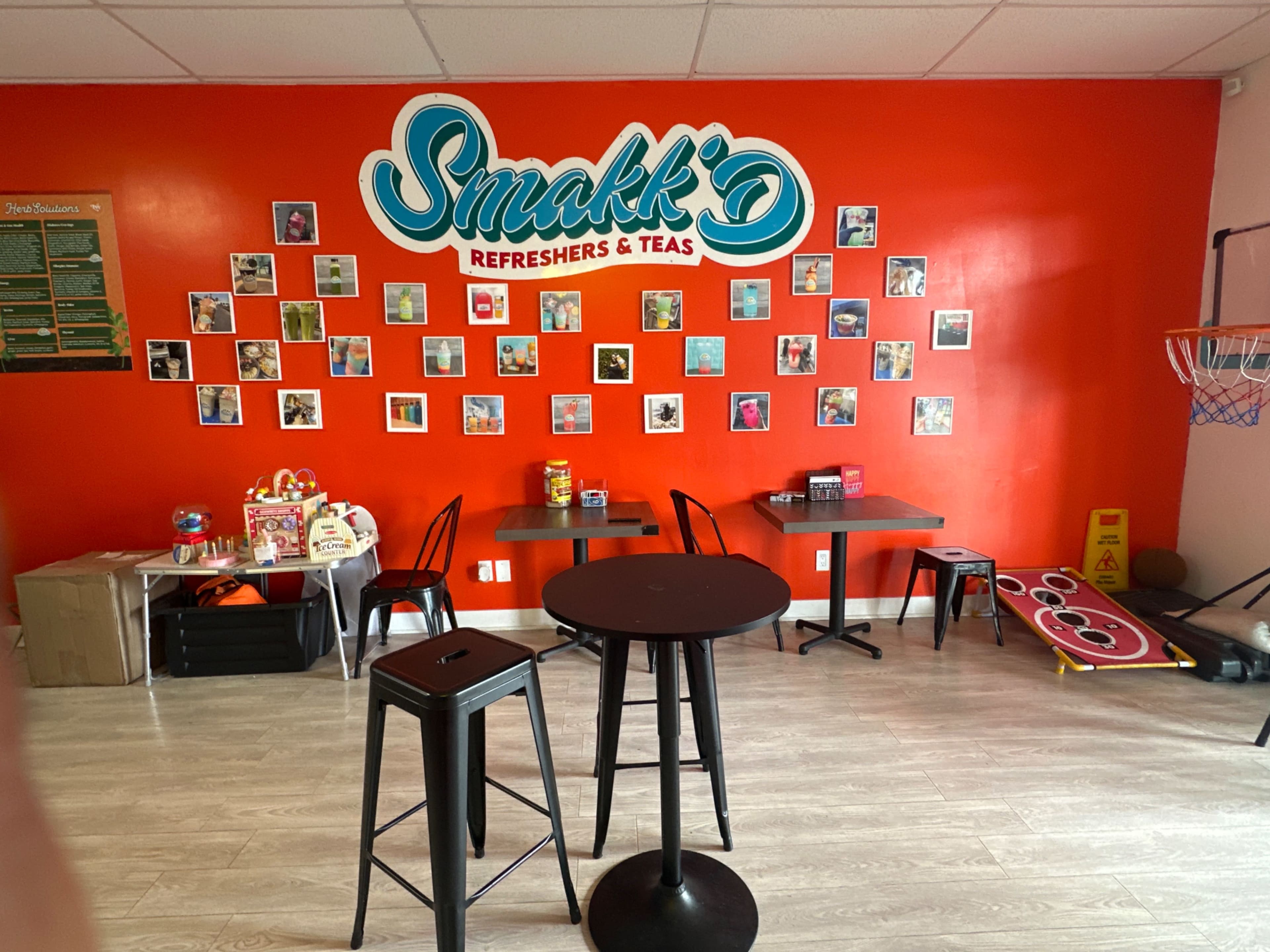 The image shows a brightly colored cafe interior with a red wall displaying a variety of framed photos, two small tables, and a basketball hoop in the corner.