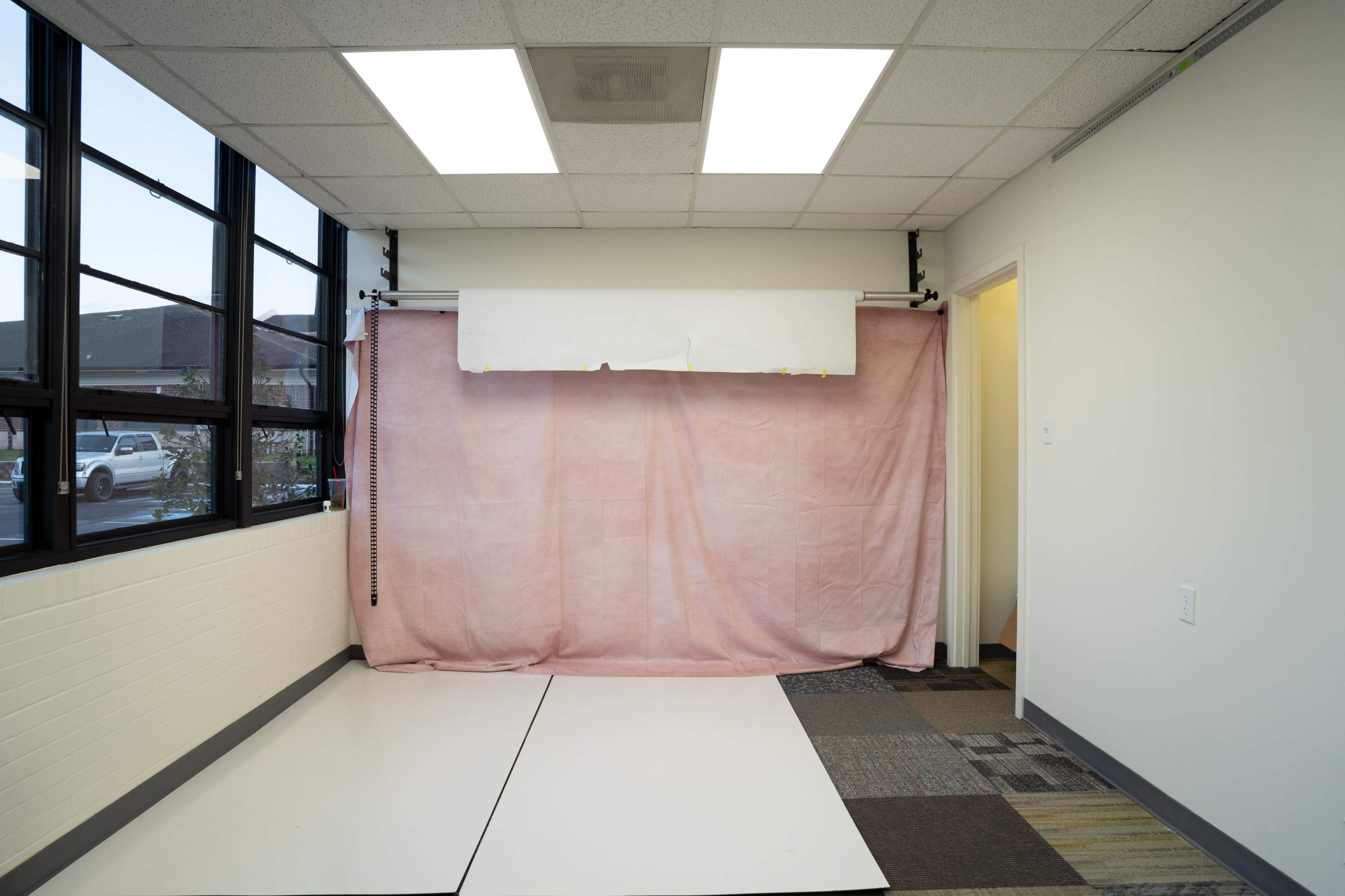 The image shows a sparsely furnished room with a pink curtain backdrop, white ceiling tiles, and mixed flooring.