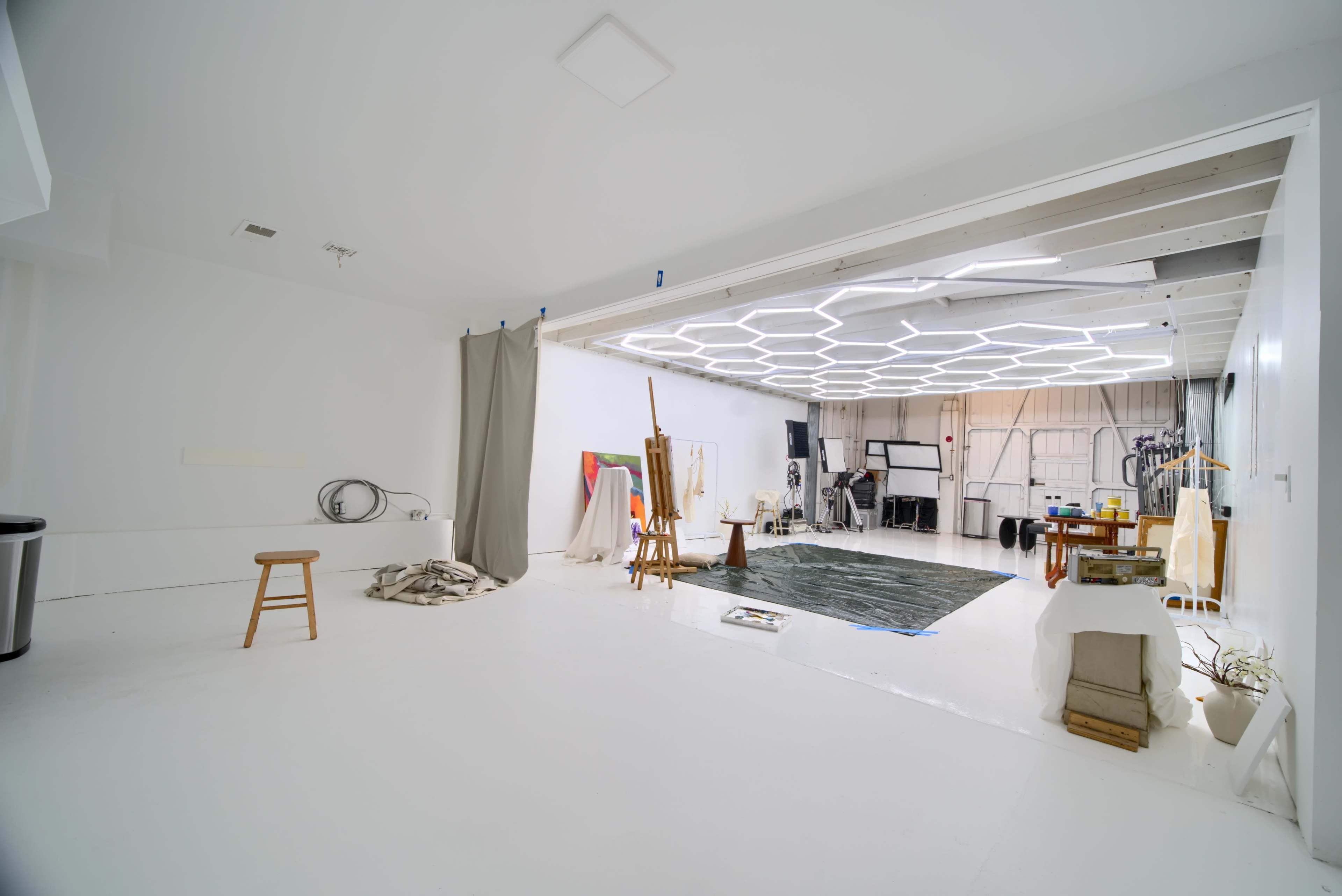 The image shows a spacious white studio with a variety of art supplies and furniture, including a stool, a large rug, and easels, illuminated by a hexagonal ceiling light.