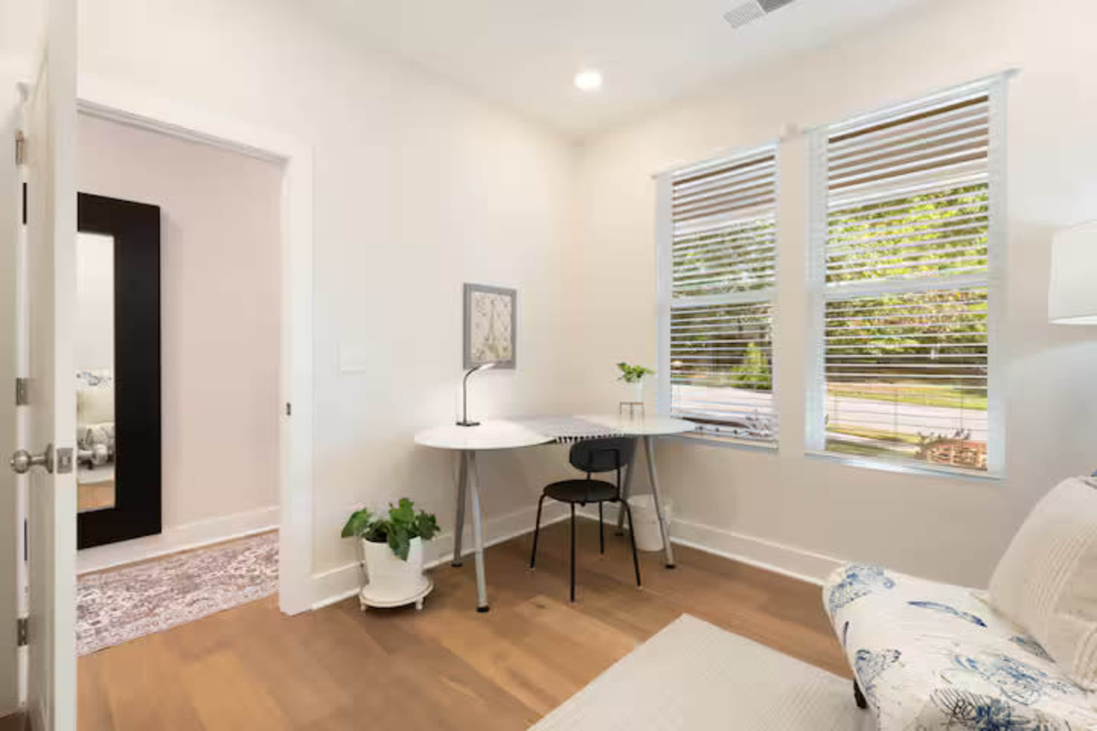 The image shows a small, well-lit home office area with a desk, a chair, and a large mirror adjacent to a doorway.