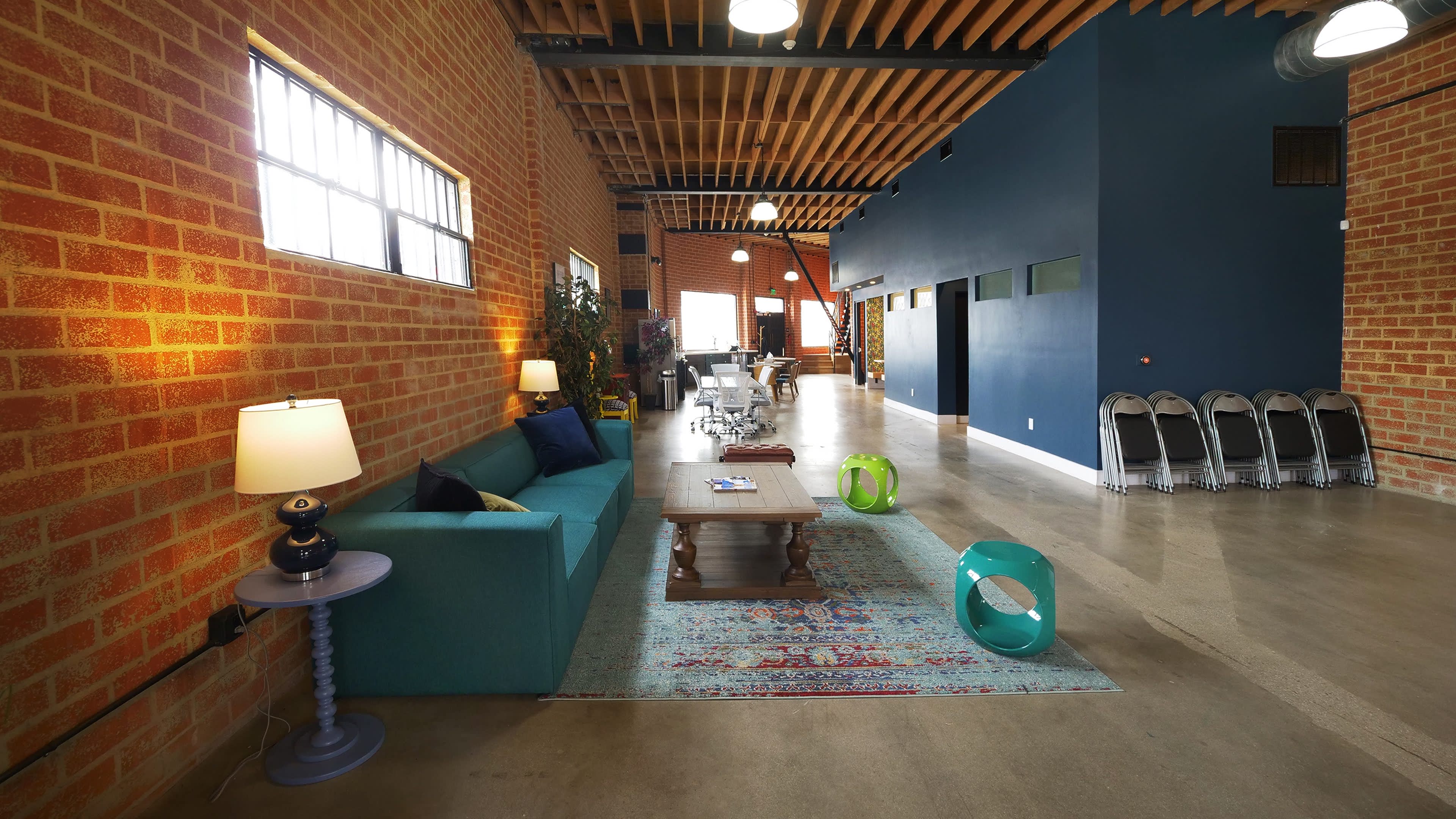 The image shows a spacious interior with exposed brick walls, modern furnishings, and a mix of seating areas, including a blue sofa and a table, illuminated by overhead lights.