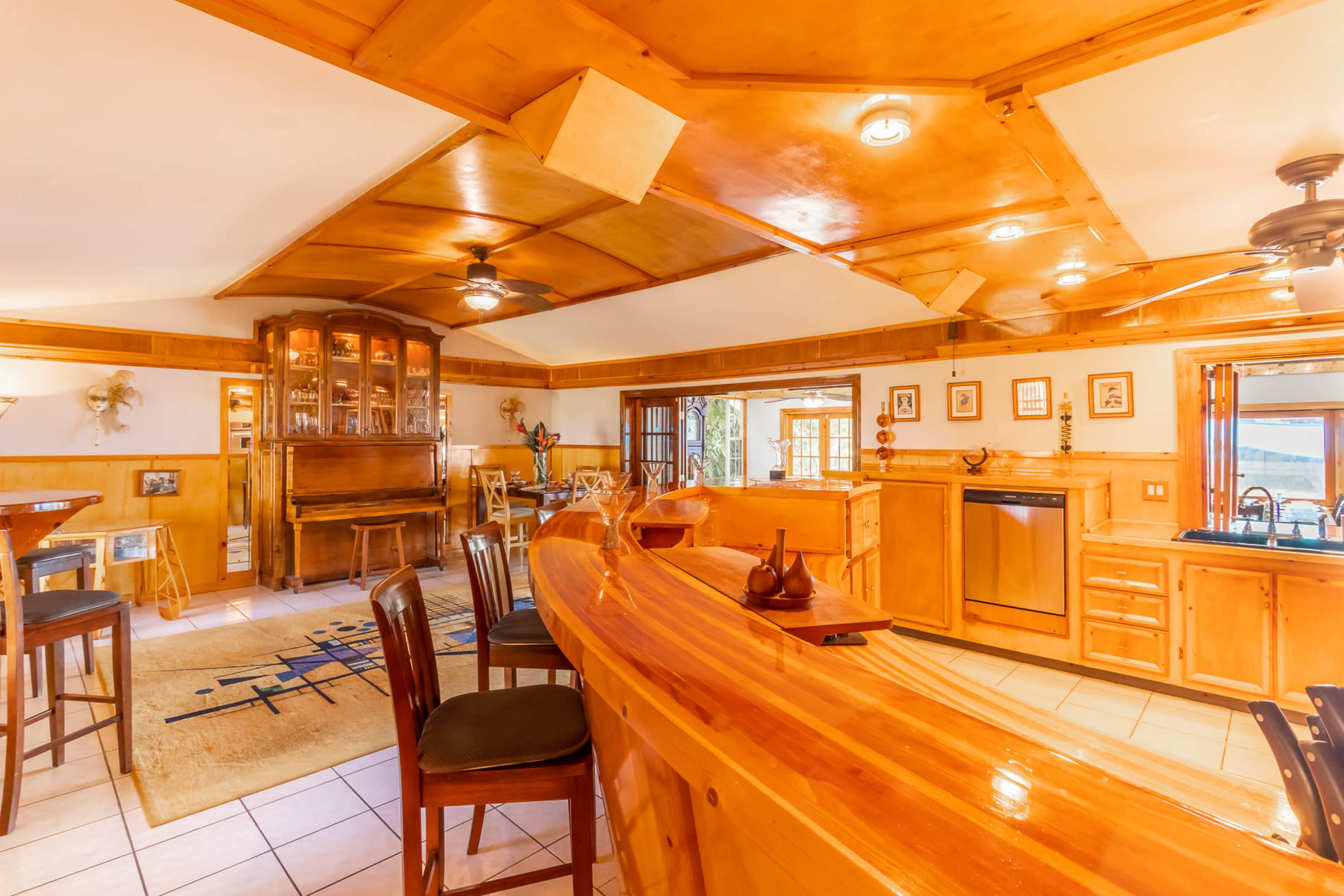 A wooden bar area with high stools, a piano in the corner, and a spacious kitchen featuring cabinets and a window overlooking a scenic view.