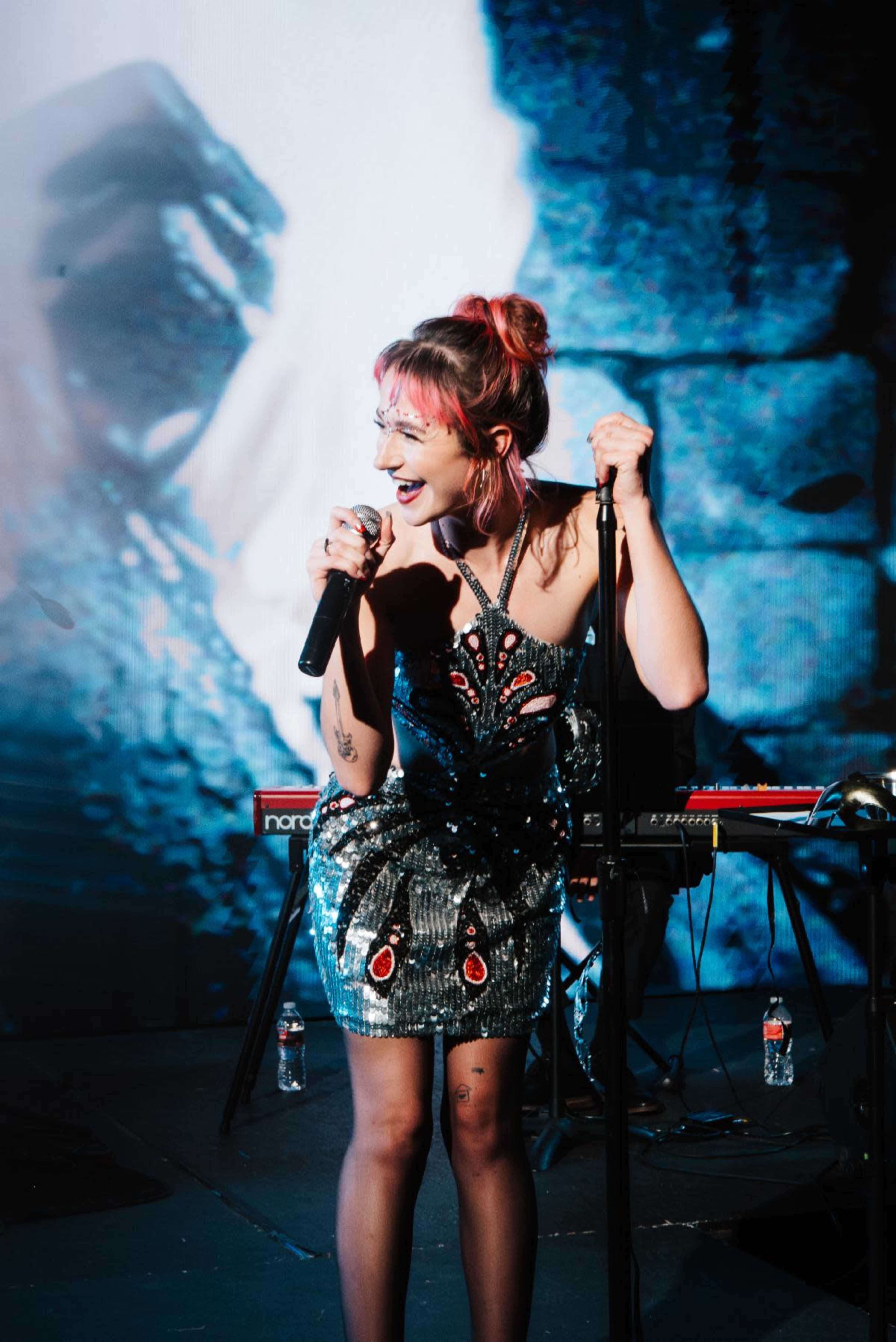 A performer sings into a microphone on stage, wearing a sequined dress, with a keyboard and water bottles visible in the background.