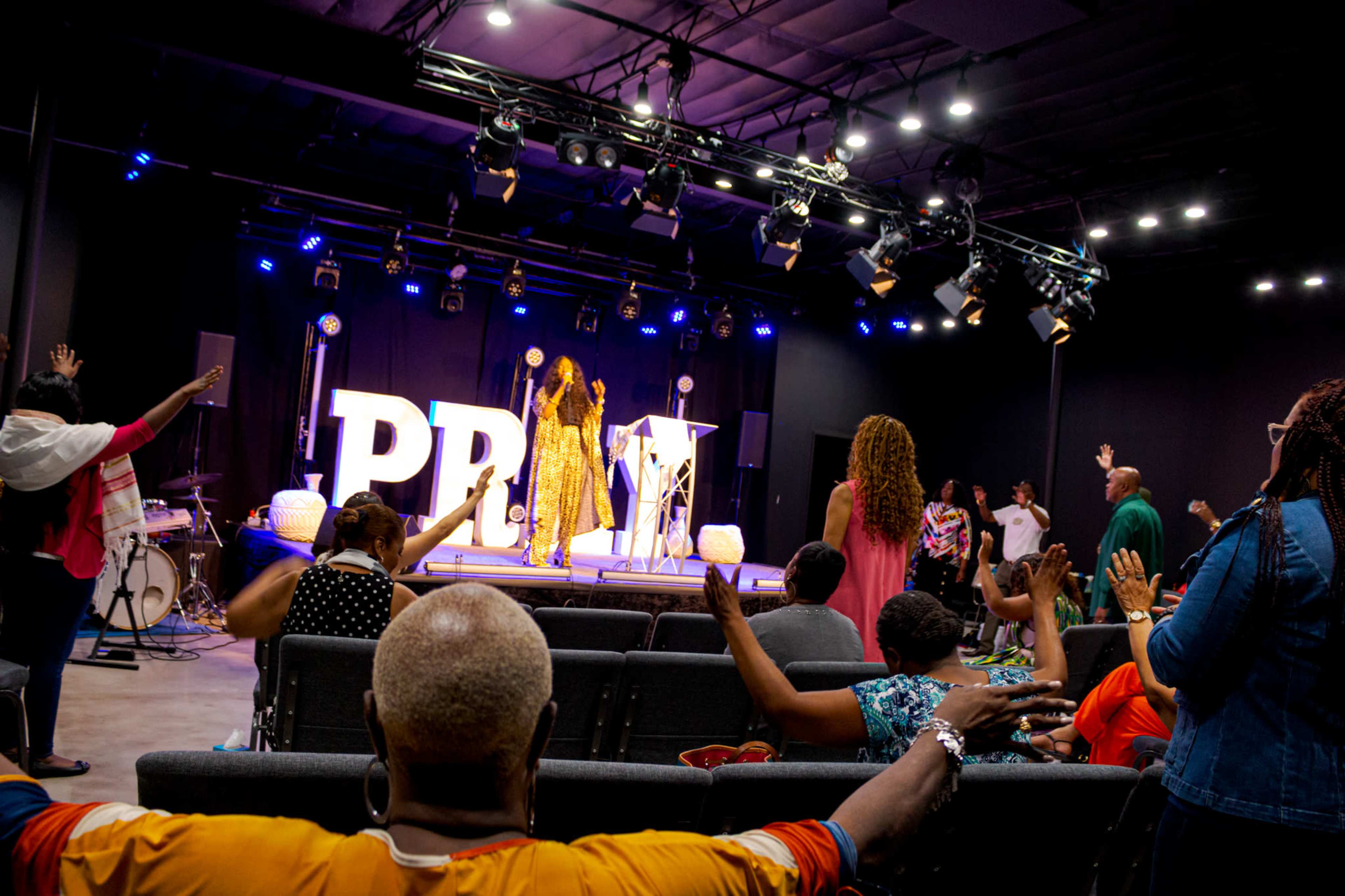 A group of people worships in a lit auditorium while a speaker addresses the audience from the stage.