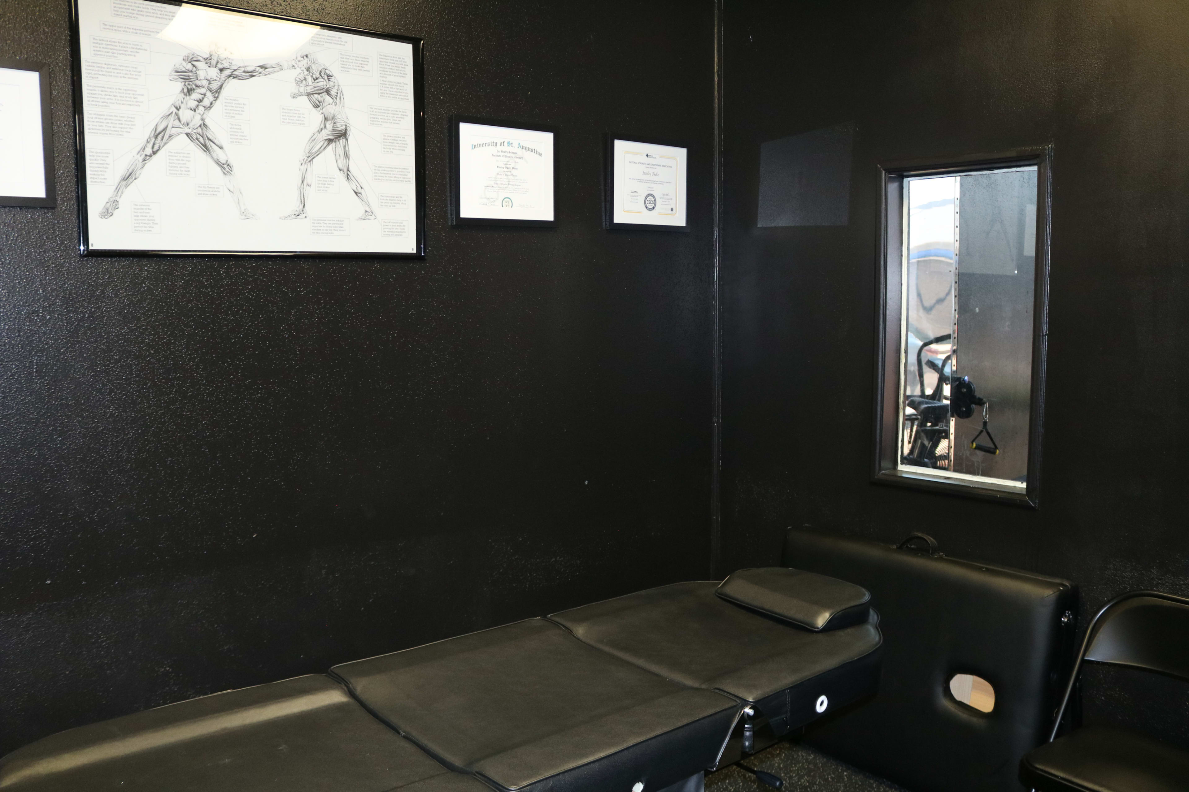 A dimly lit room with a black wall, a treatment table, a framed illustration of two boxers, and a small mirror.