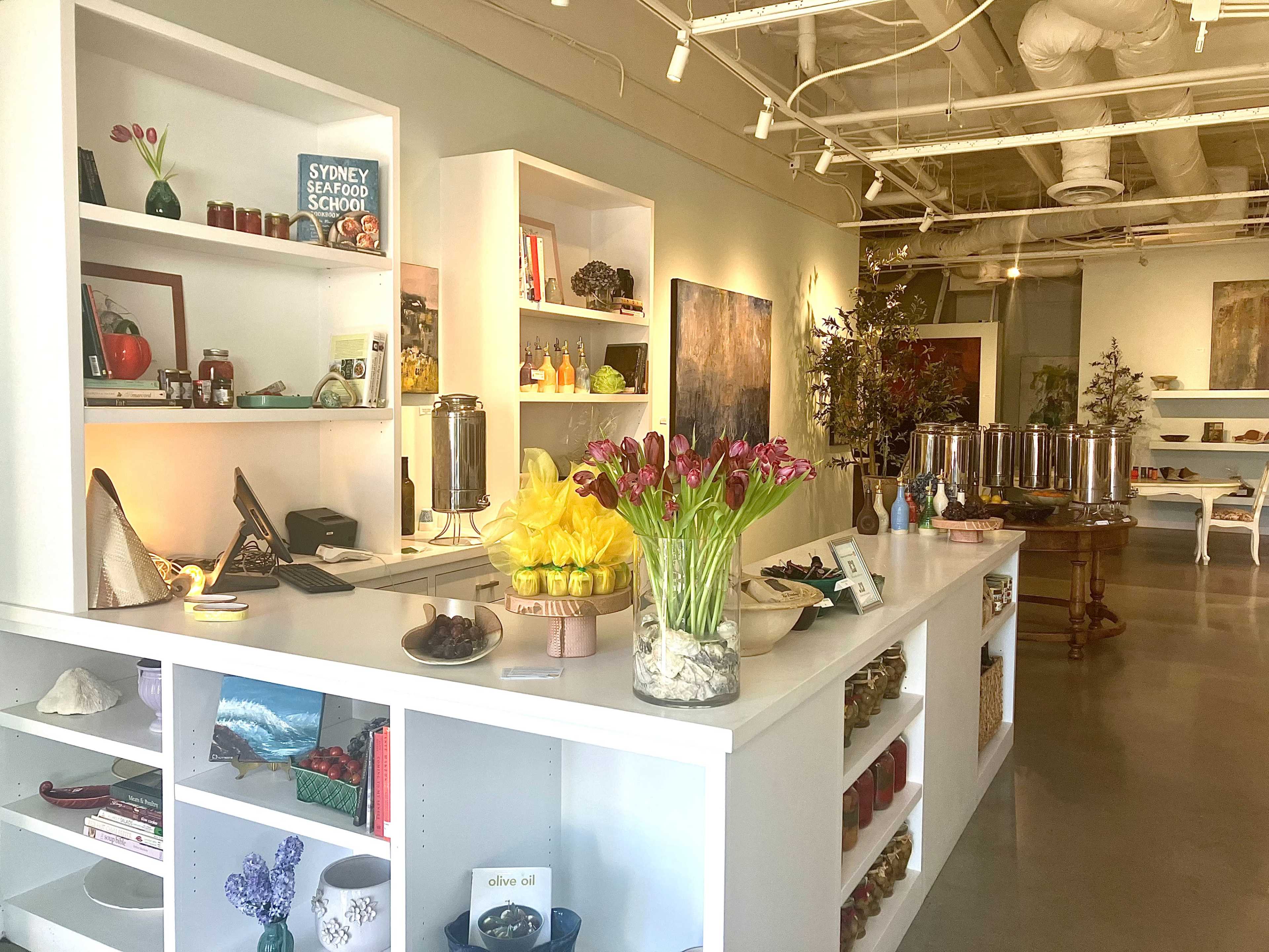 The image shows a bright, modern retail space featuring shelves filled with various food items, decorative objects, and a bouquet of tulips on a counter.