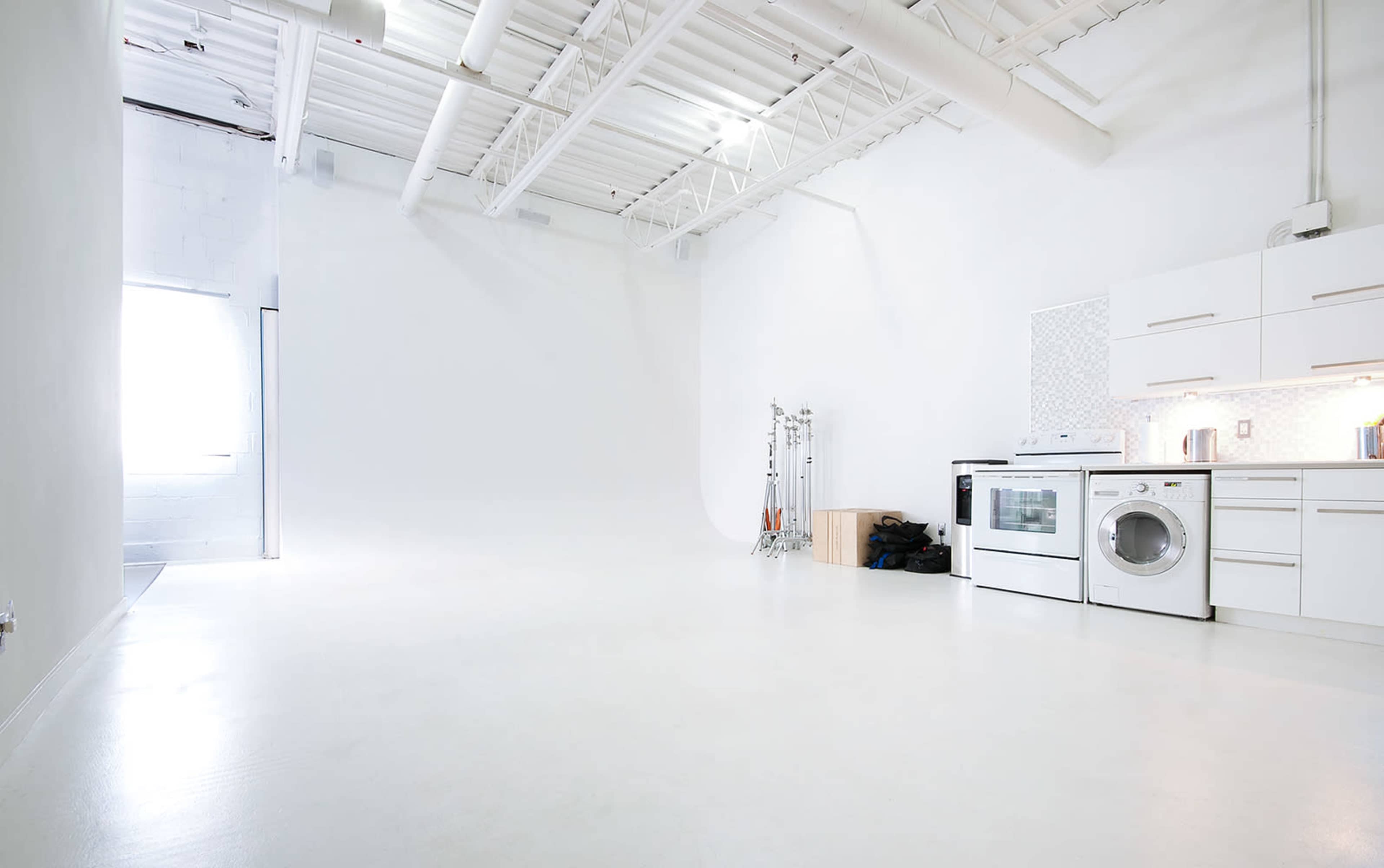 The image shows a spacious, bright studio with white walls and a polished floor, featuring a kitchen area with a stove and washing machine in the corner.