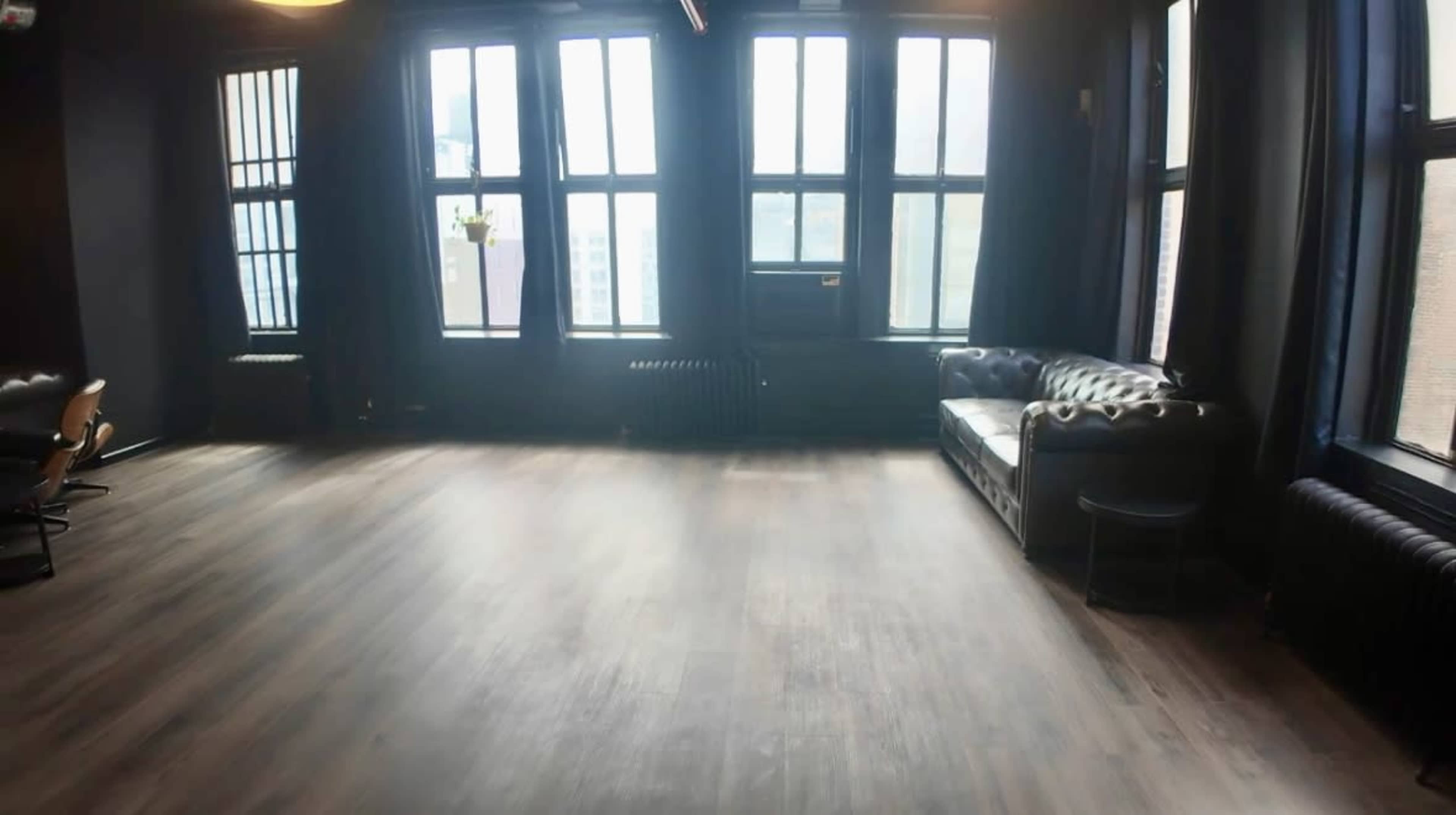 The image shows a spacious room with large windows, hardwood floors, and a black leather couch along one wall.