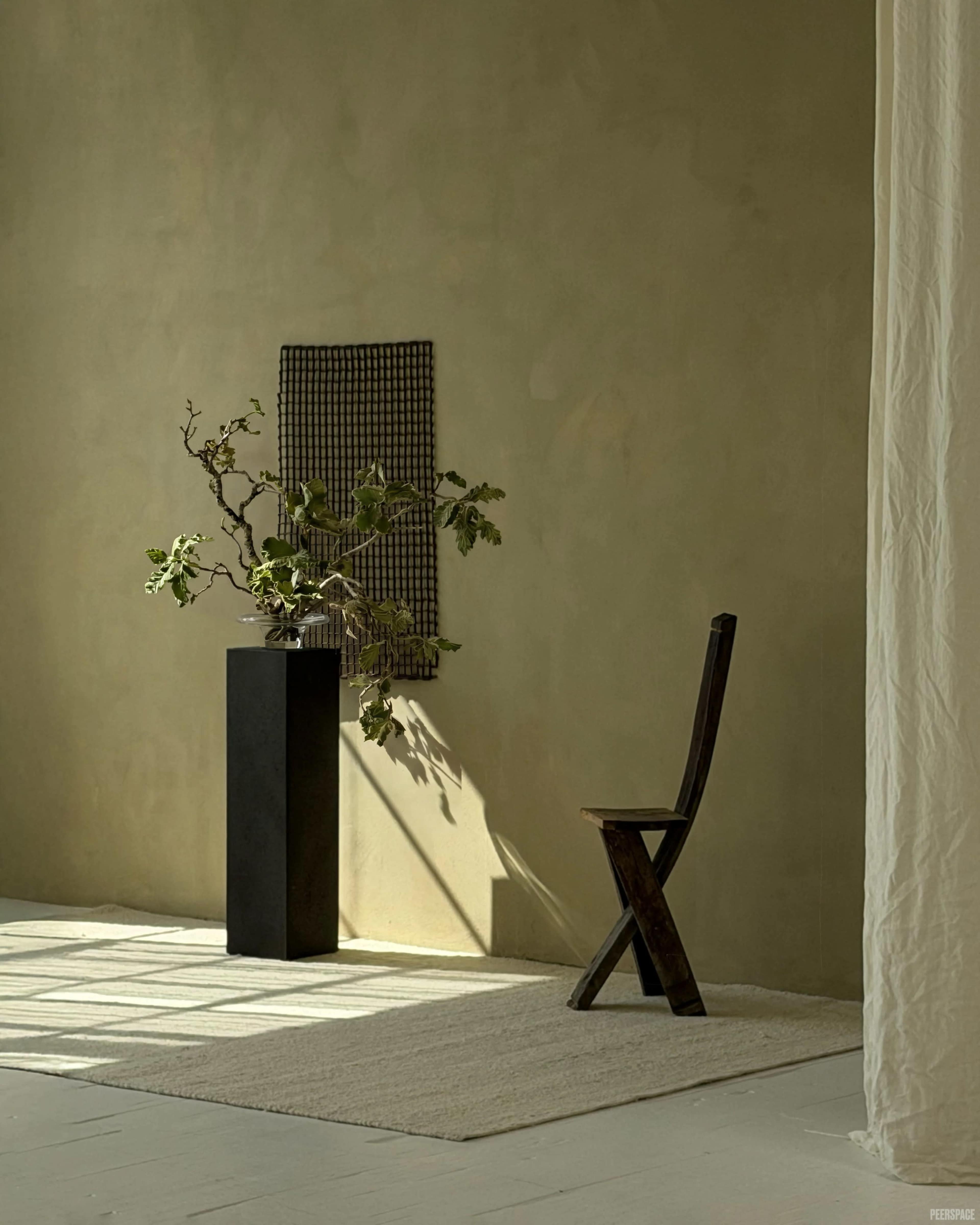 The image features a minimalist interior with a wooden chair next to a black pedestal displaying a floral arrangement, against a soft beige wall.