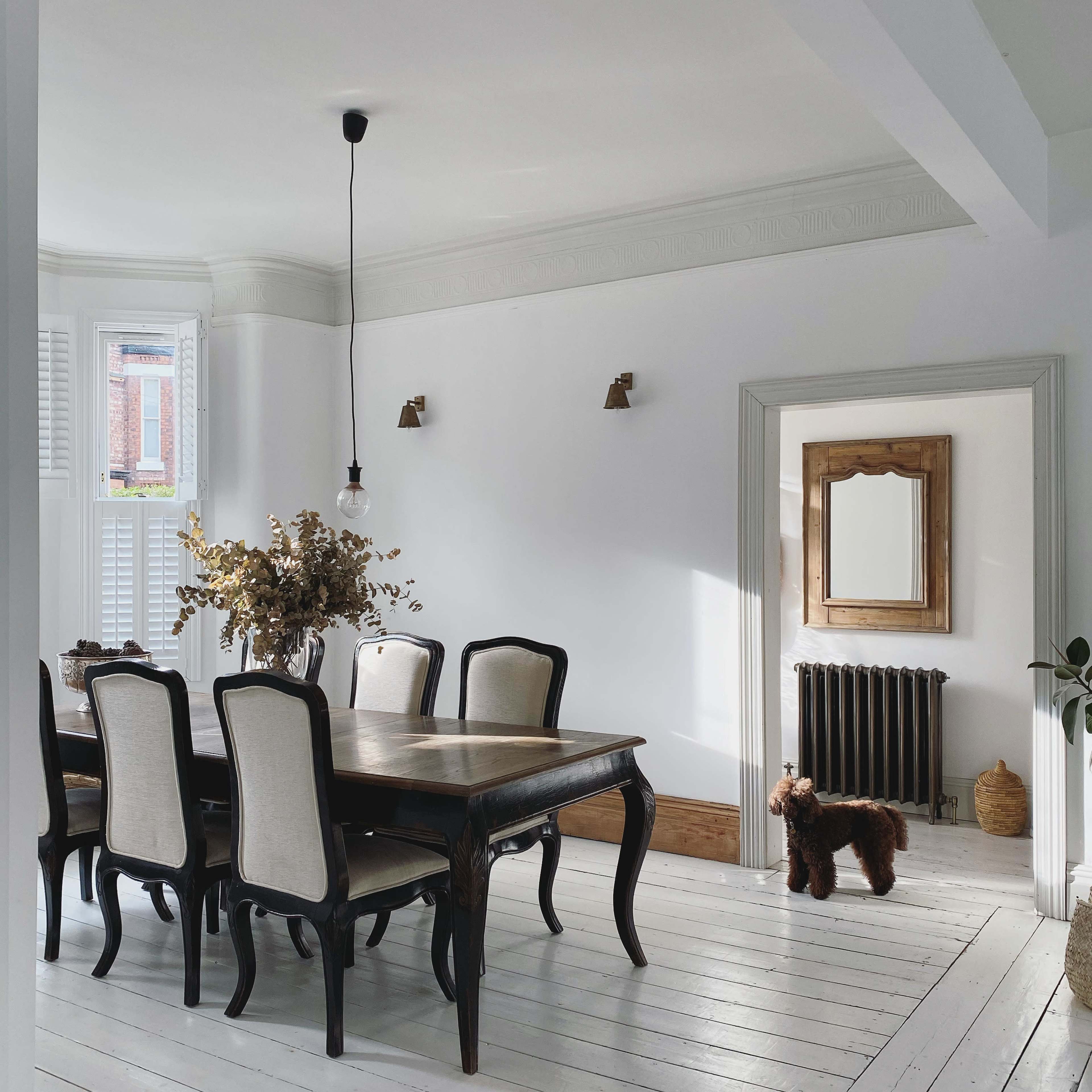 A dining room features a wooden table with six chairs, a vase of dried flowers, and a small dog standing near a door.