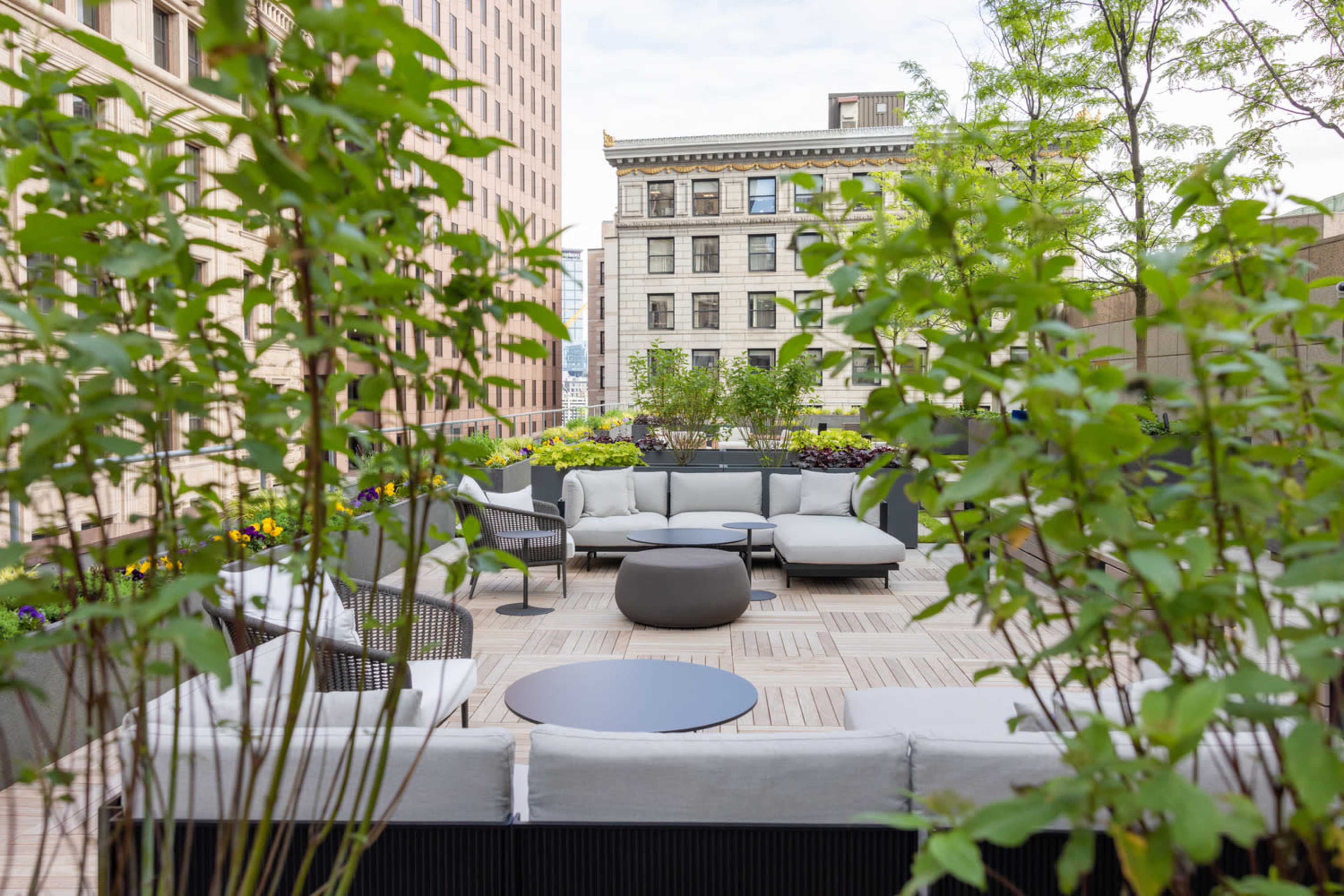 A modern patio area with seating arrangements surrounded by greenery and flowers, set in an urban environment.
