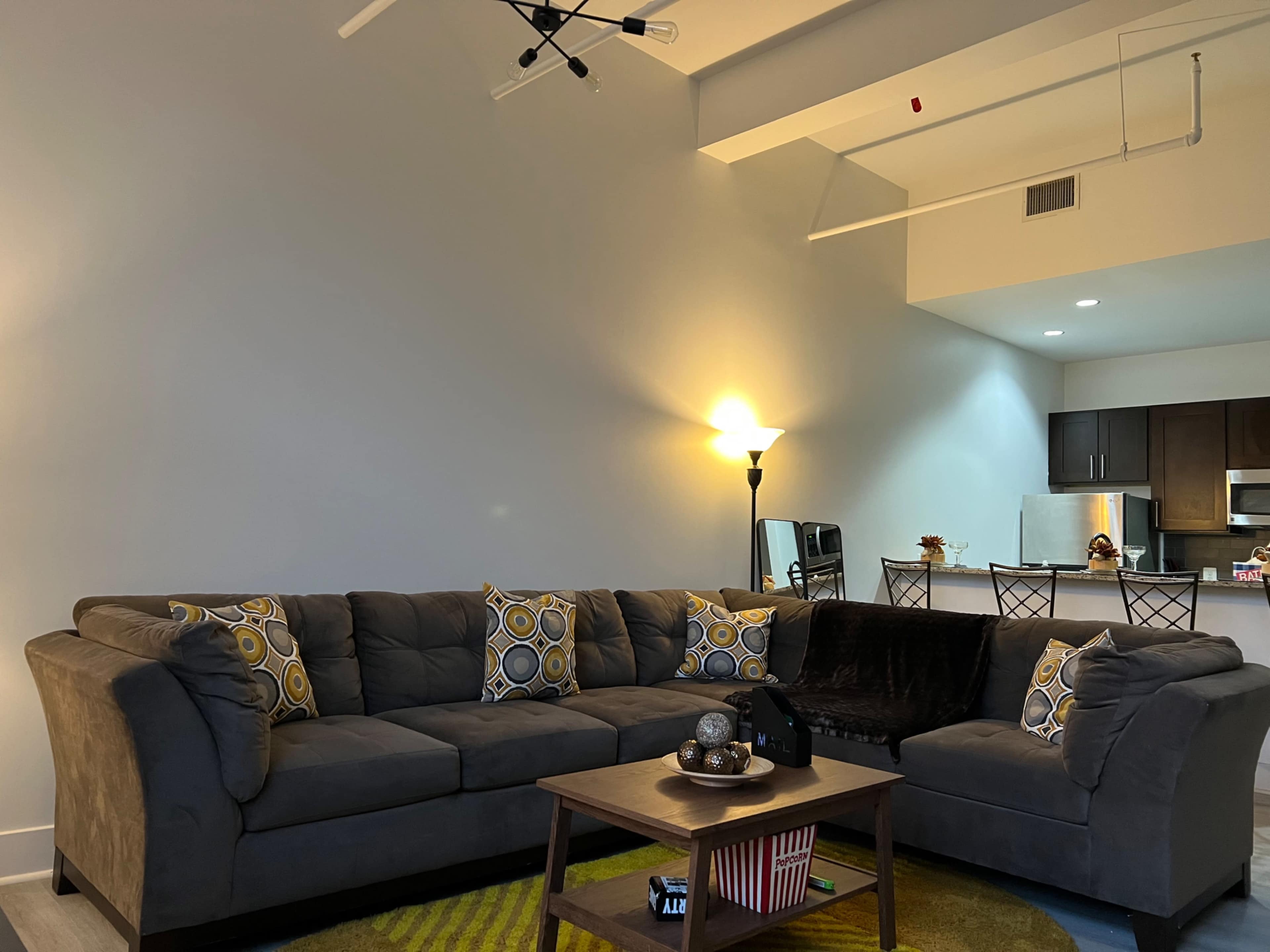 The image shows a modern living room with a sectional gray sofa, a coffee table, and a kitchen area in the background.