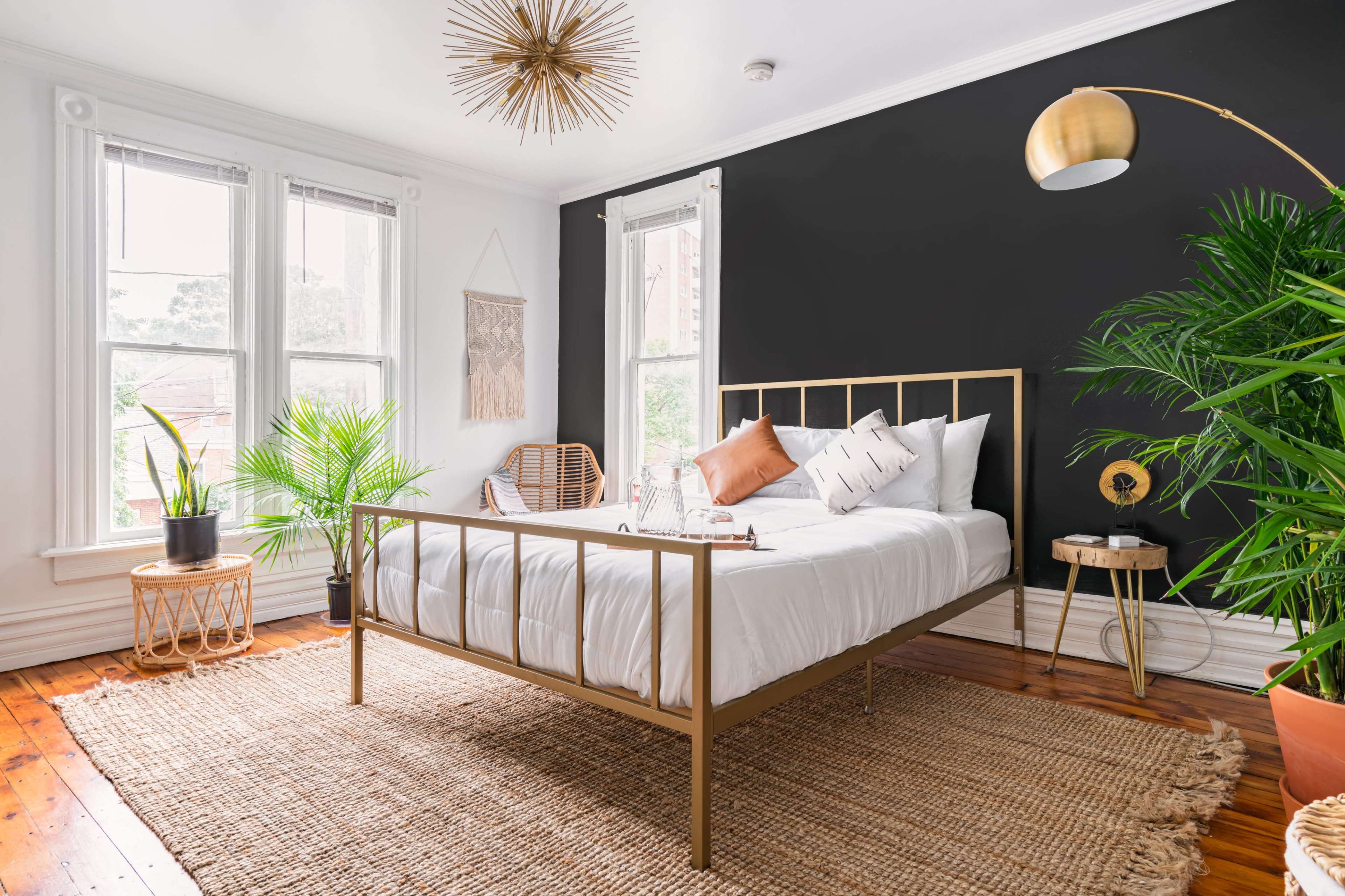 A modern bedroom featuring a gold metal bed frame, white bedding, a rattan chair, large indoor plants, and a dark accent wall.