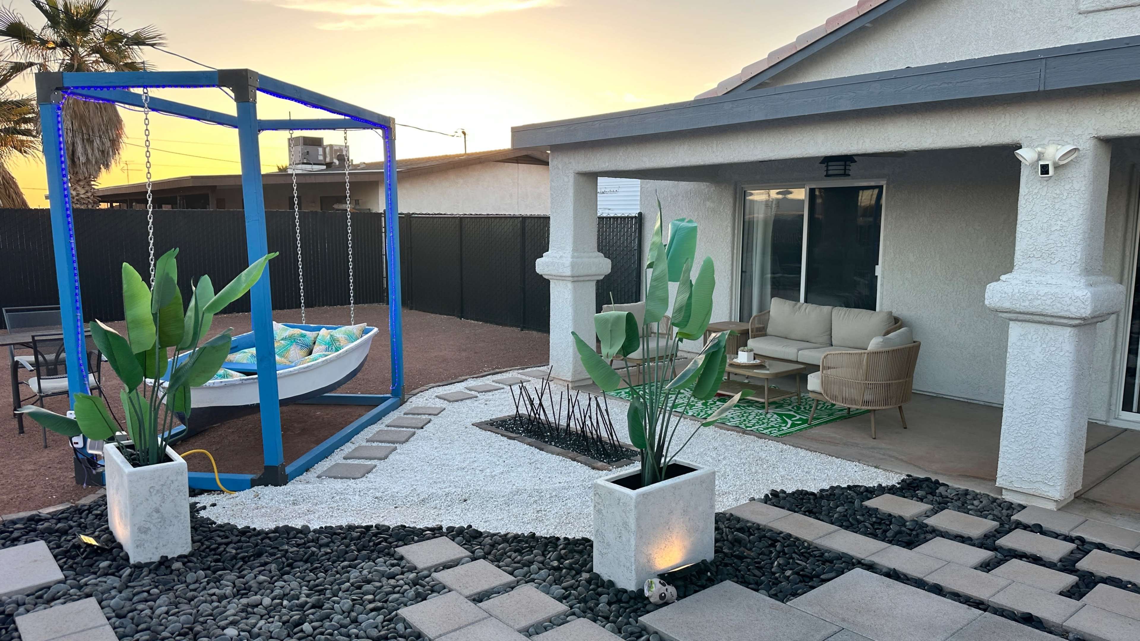 A patio area features a swing set, potted plants, and a lounge arrangement beside a gravel path at sunset.