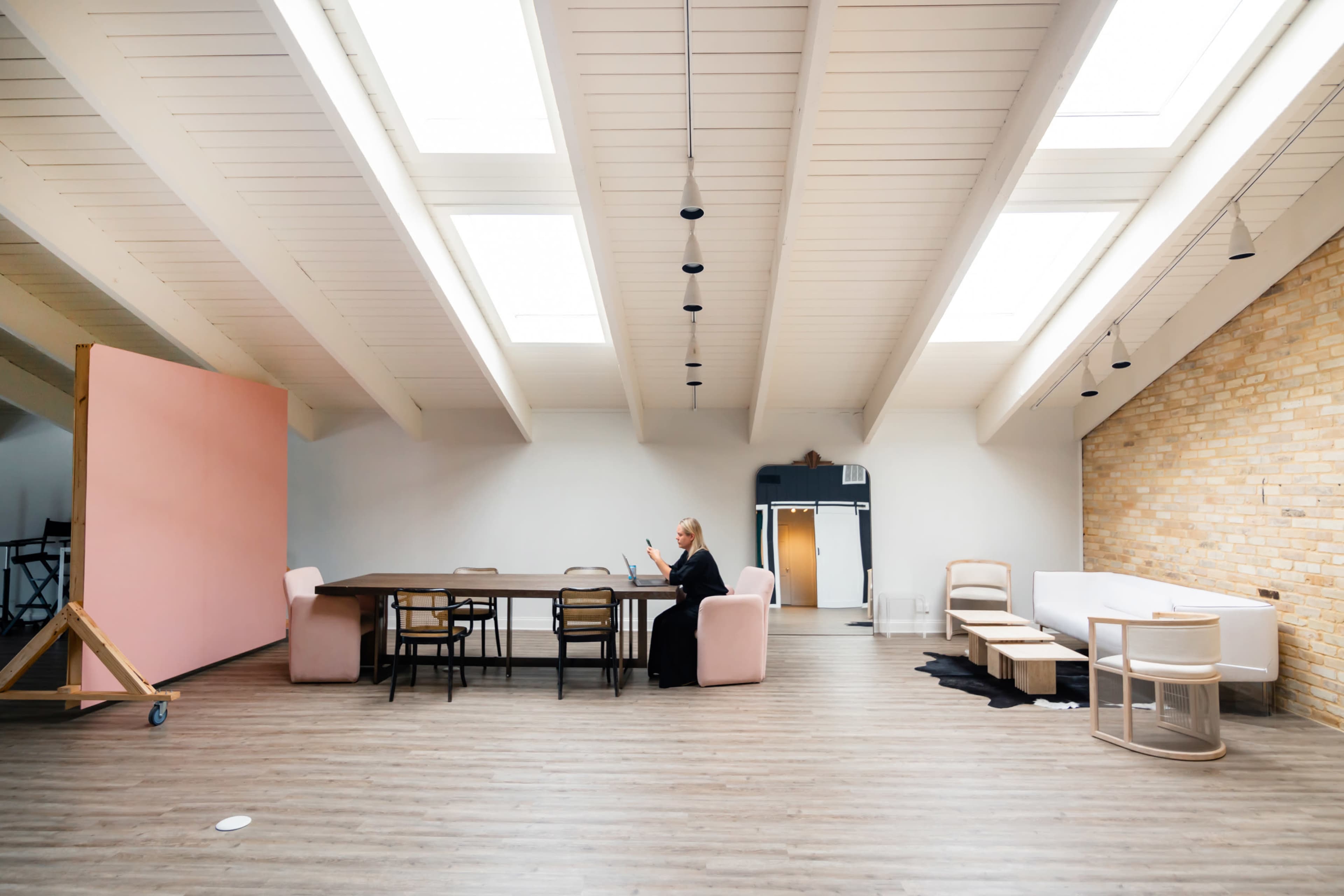 A spacious interior with a long wooden table, several chairs, and a woman sitting on a pink armchair, surrounded by large skylights and a mix of brick and painted walls.
