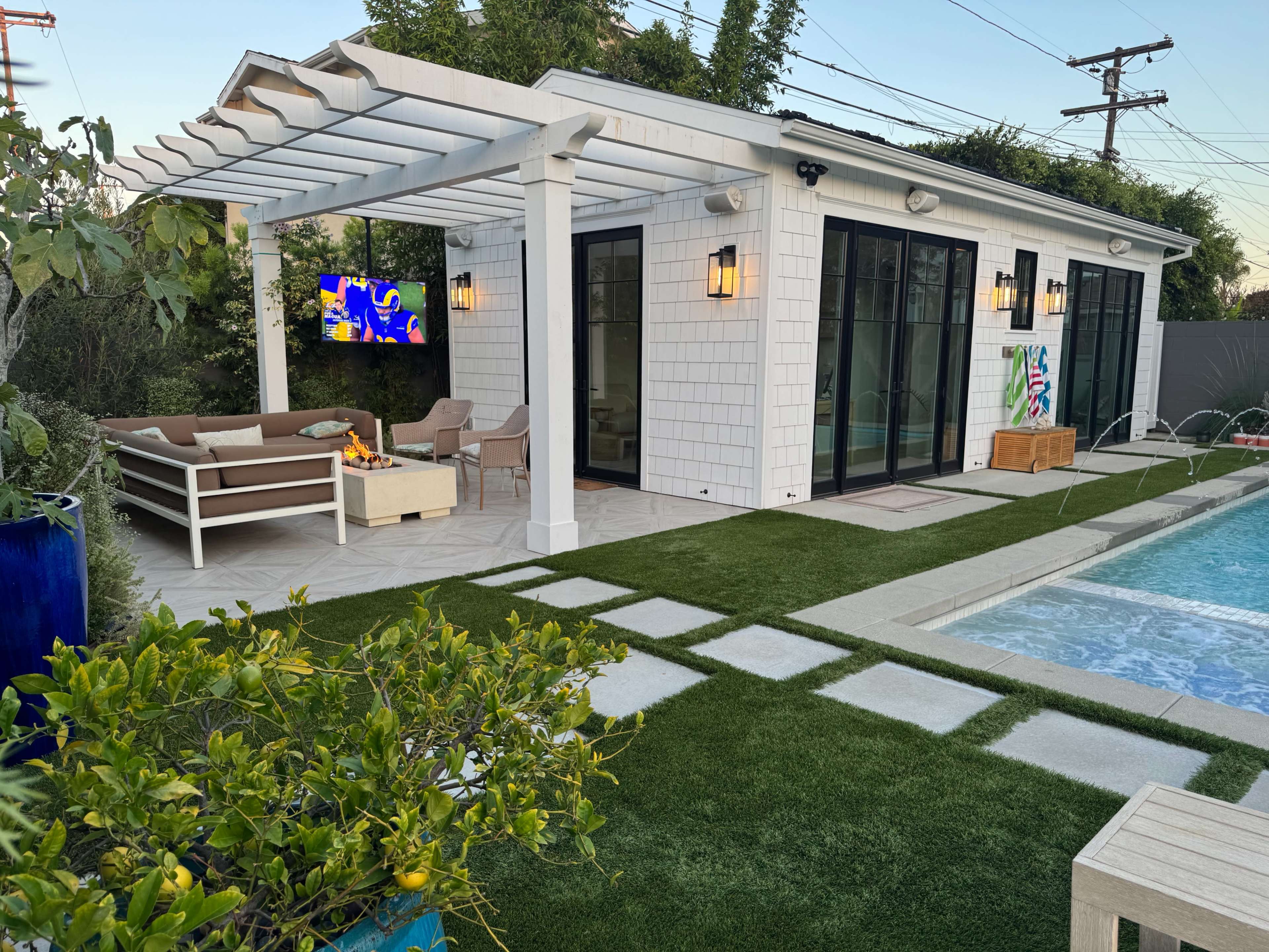 A modern outdoor living space features a pool, seating area, and a covered patio with a TV and fire pit.