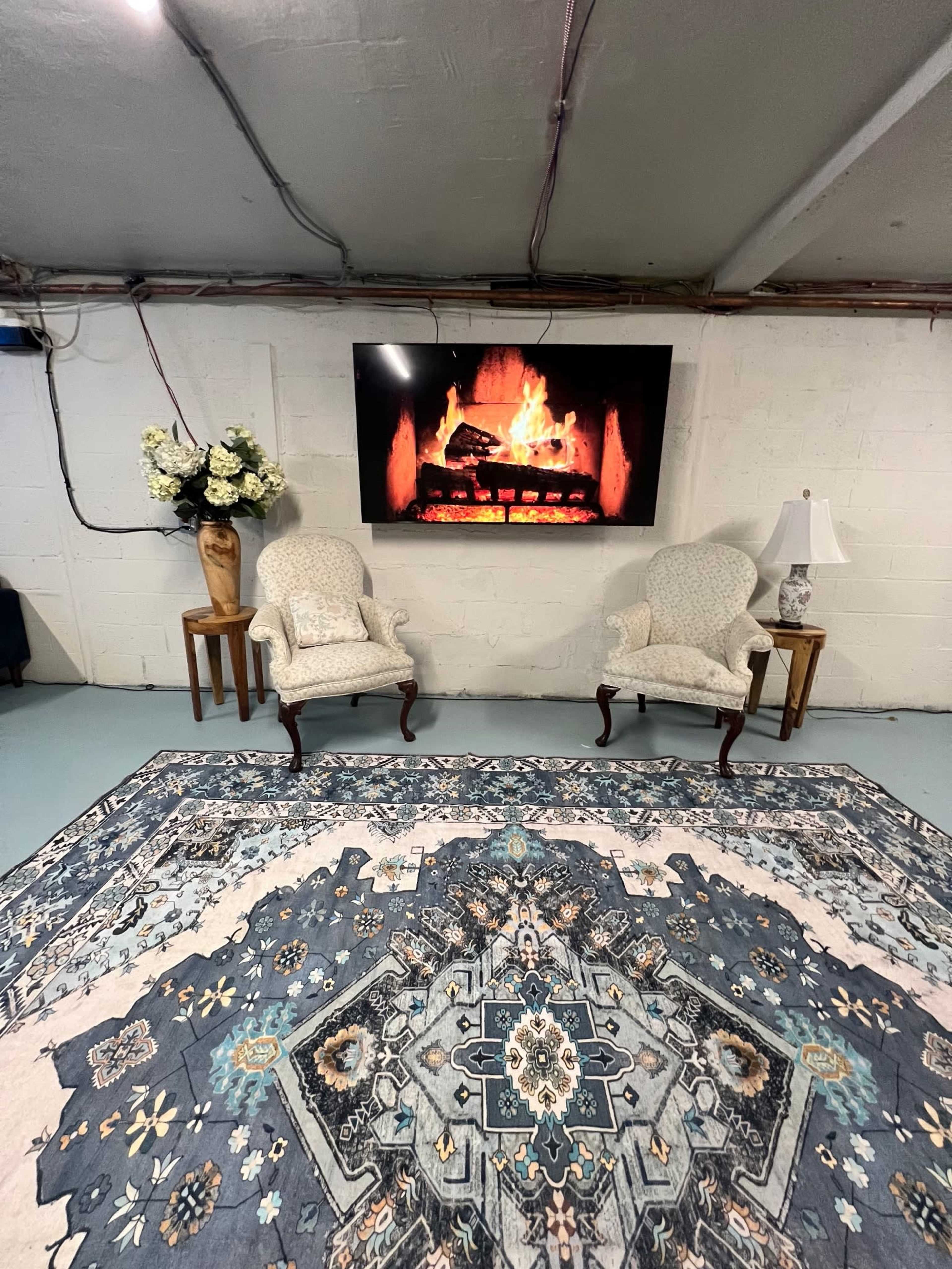 The image shows two upholstered chairs placed in front of a framed digital fireplace, with a floral arrangement on a pedestal and a lamp on a side table, all situated on an ornate area rug in a basement setting.