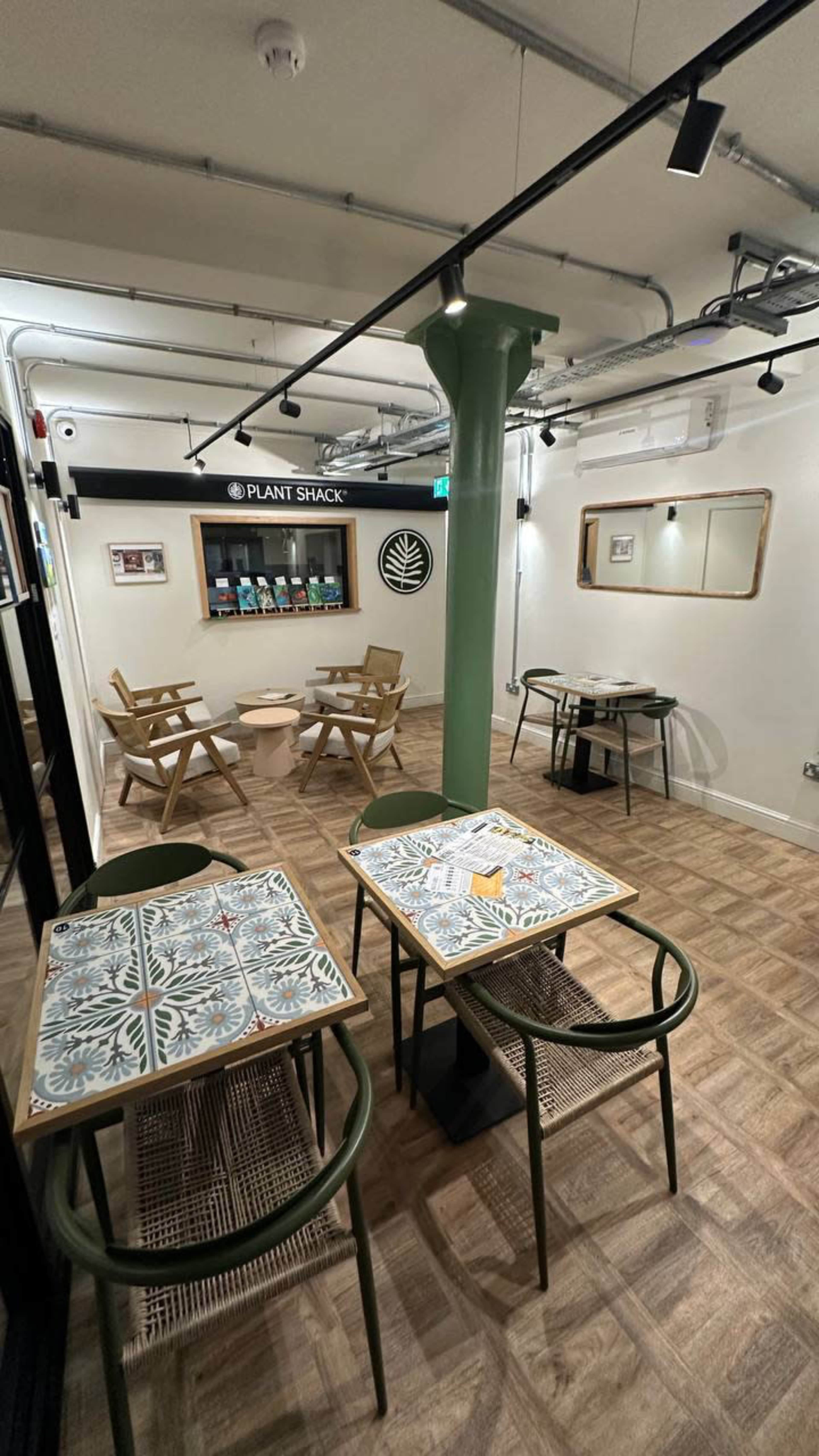 The image shows a modern café interior with wooden tables and chairs, featuring decorative tiles and a green support pillar in the center.