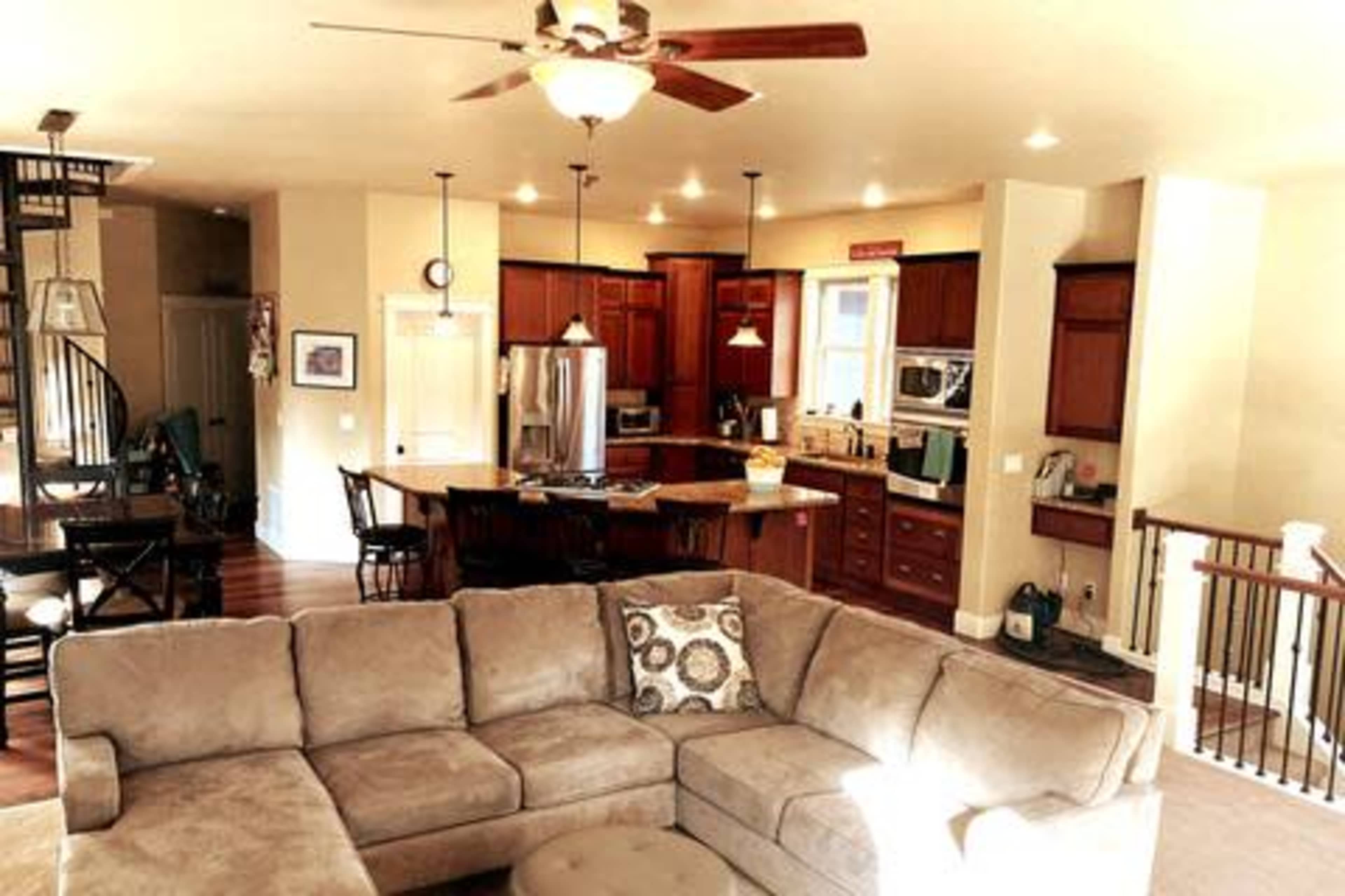 An open-concept living space featuring a beige sectional sofa in the foreground, a kitchen with wooden cabinets and stainless steel appliances in the background, and a dining area visible to the left.
