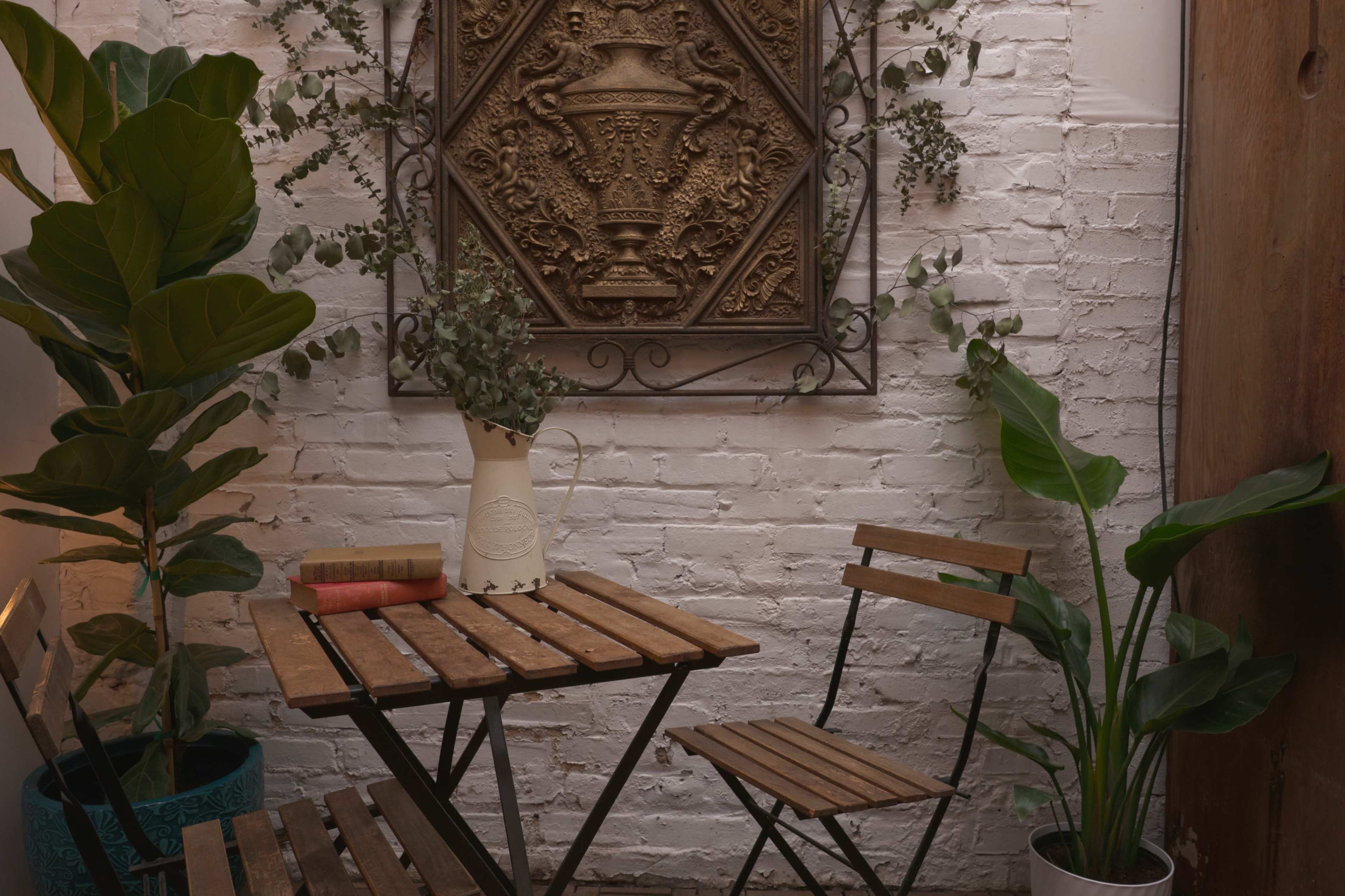 A small indoor seating area features a wooden table with two chairs, surrounded by potted plants and a decorative wall piece.