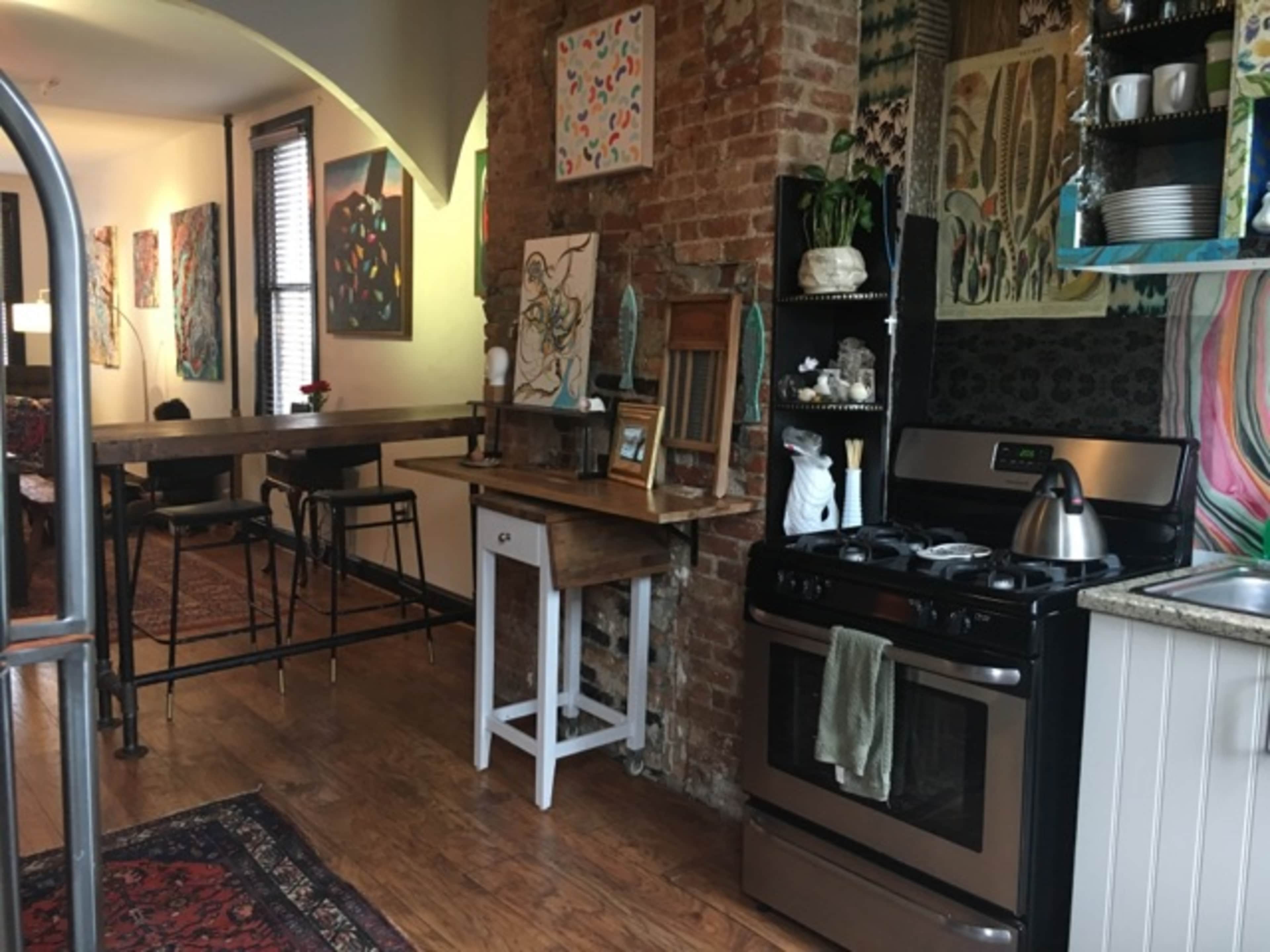 The image shows a cozy, open kitchen area with brick walls, a wooden dining bar, and various colorful artworks displayed on the walls.