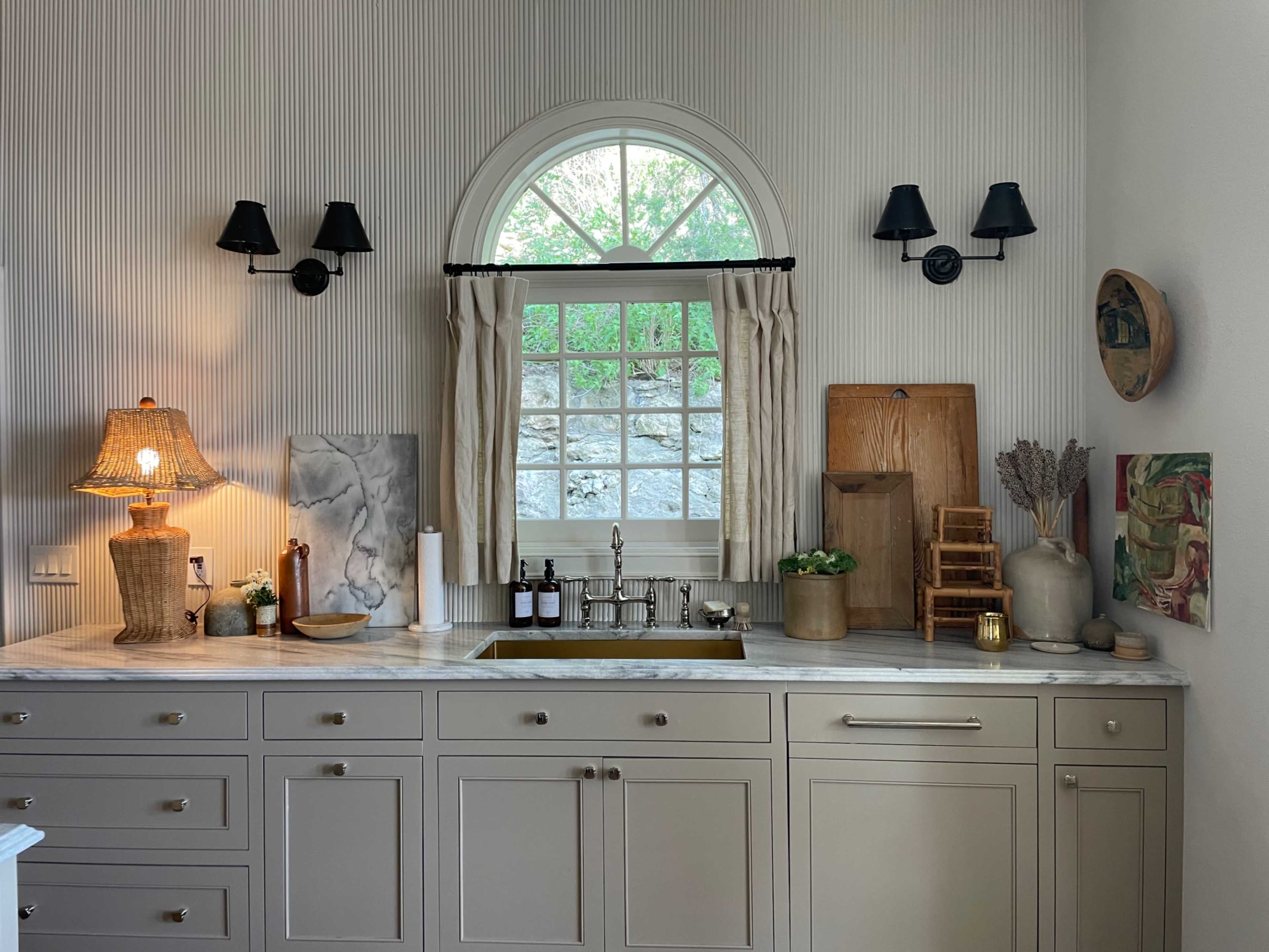 A kitchen features a marble countertop with a sink, a window dressed in curtains, and various decorative items including a lamp, paintings, and a cutting board.