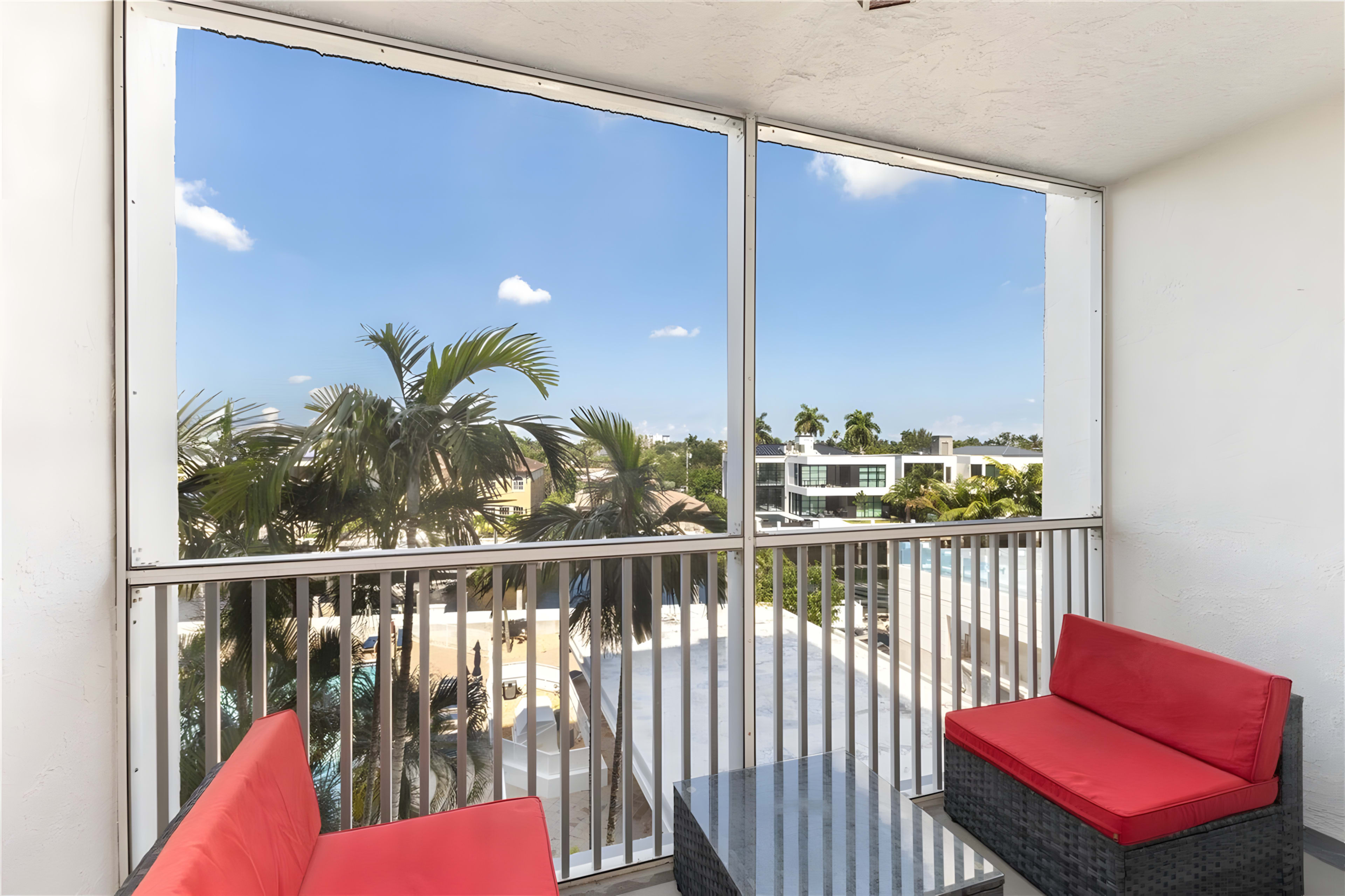The image shows a balcony with red seating and a view of a palm tree and buildings under a clear blue sky.