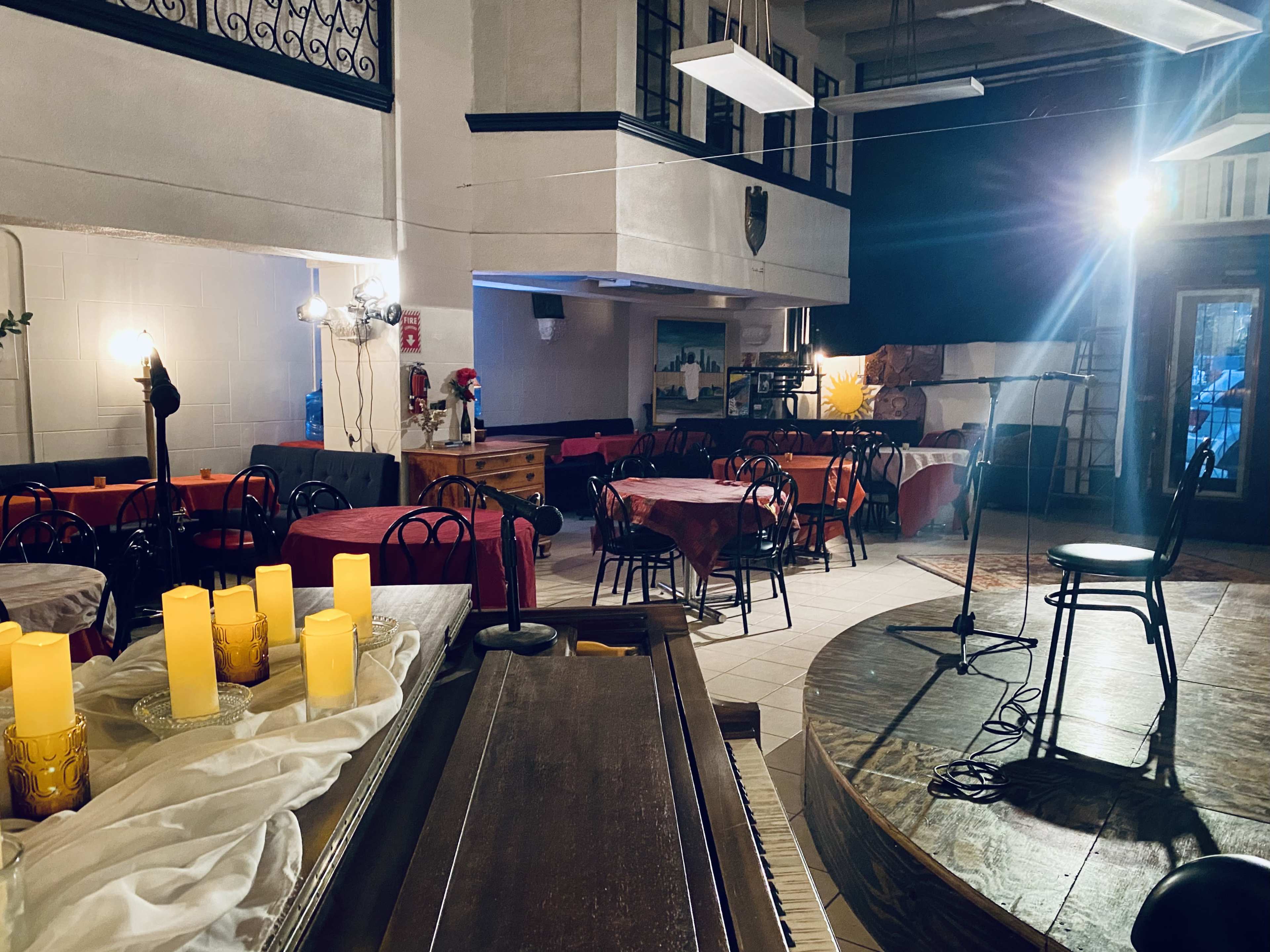 A cozy restaurant setting with tables covered in red and white linens, chairs arranged around them, and a small stage with a microphone in the corner.