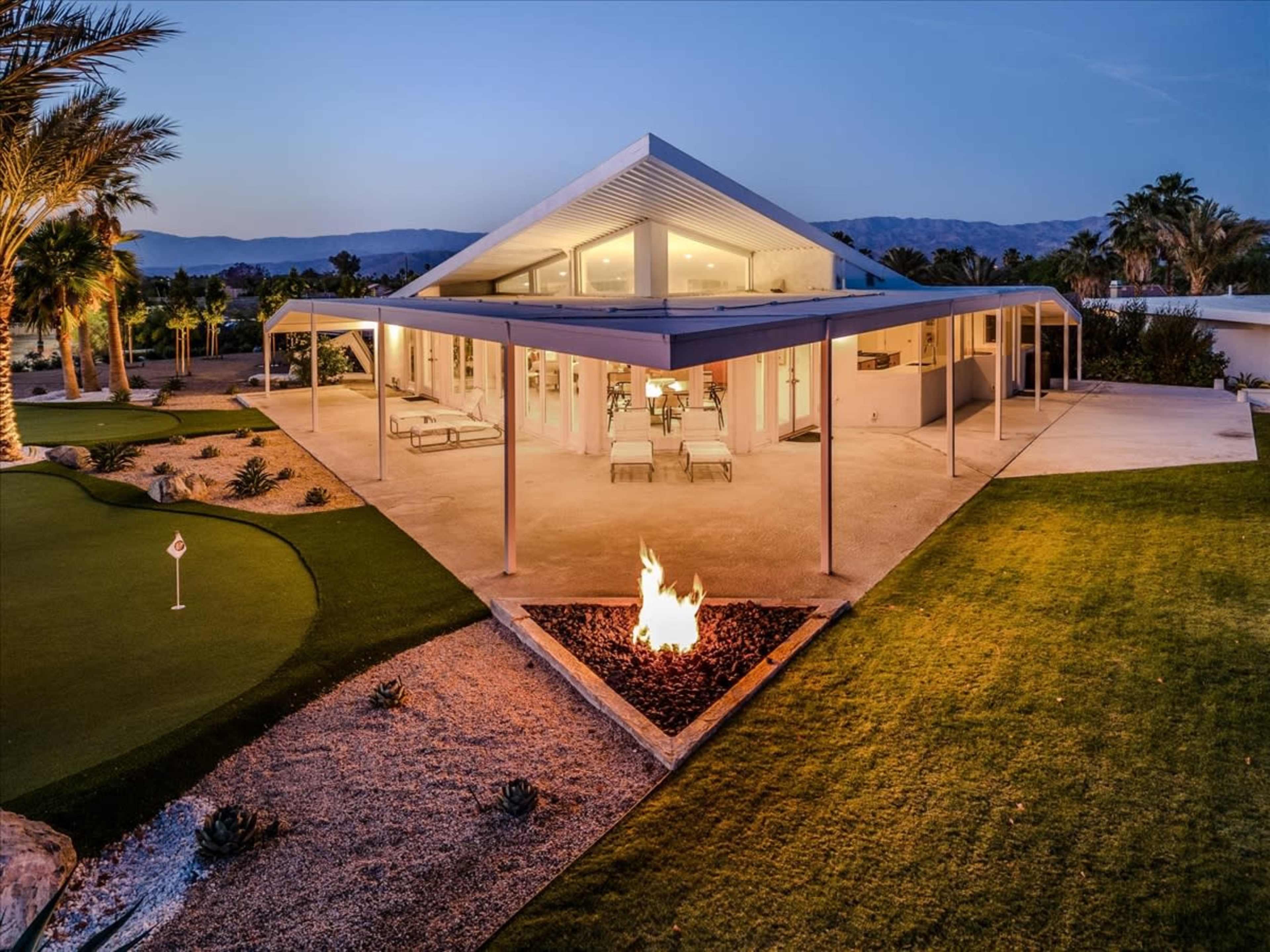 A modern house with a triangular roof and large windows is surrounded by landscaped gardens and features a fire pit in the foreground.