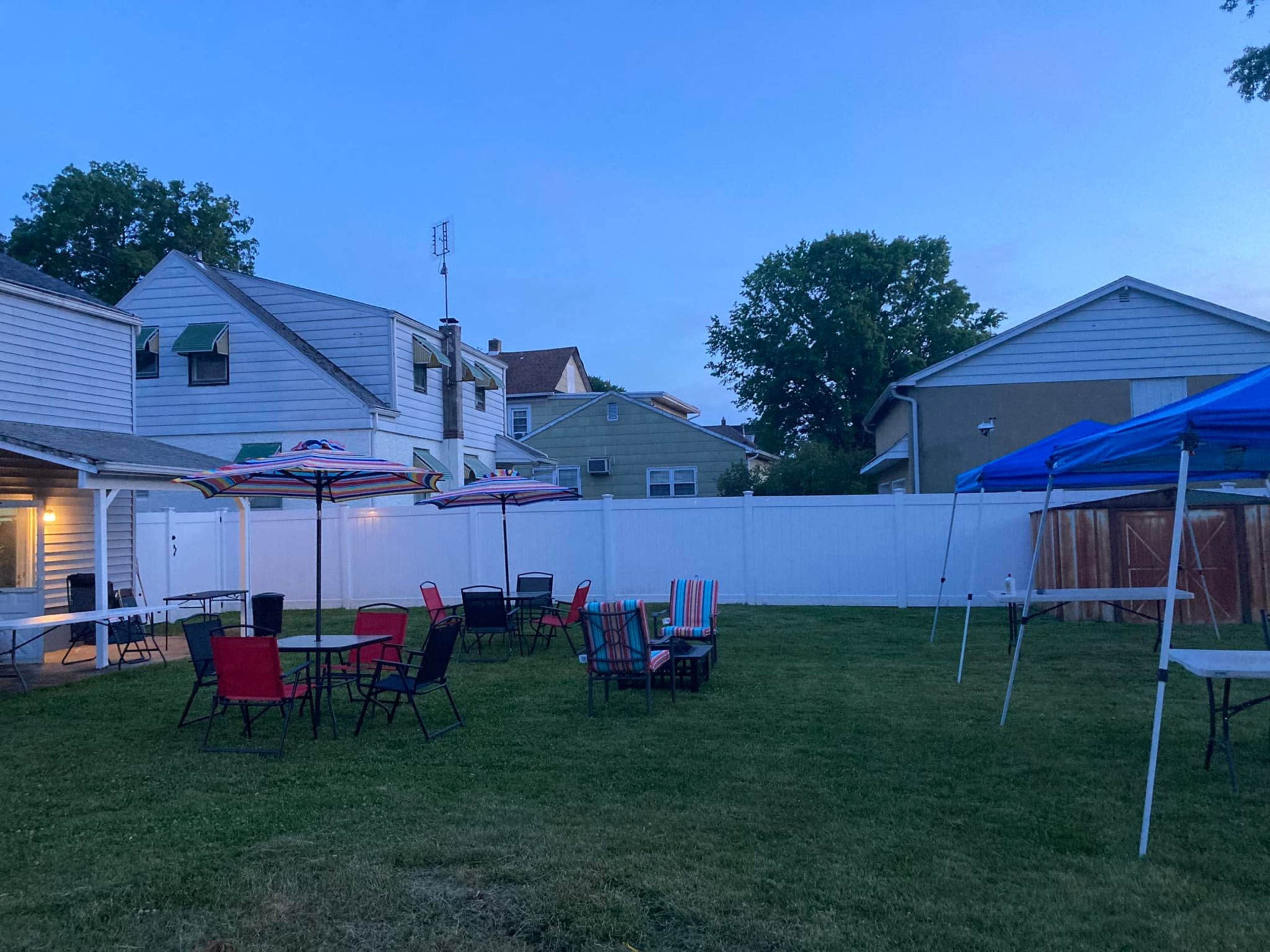 A backyard space features several tables and chairs under umbrellas, with two tents set up nearby and a white fence enclosing the area.
