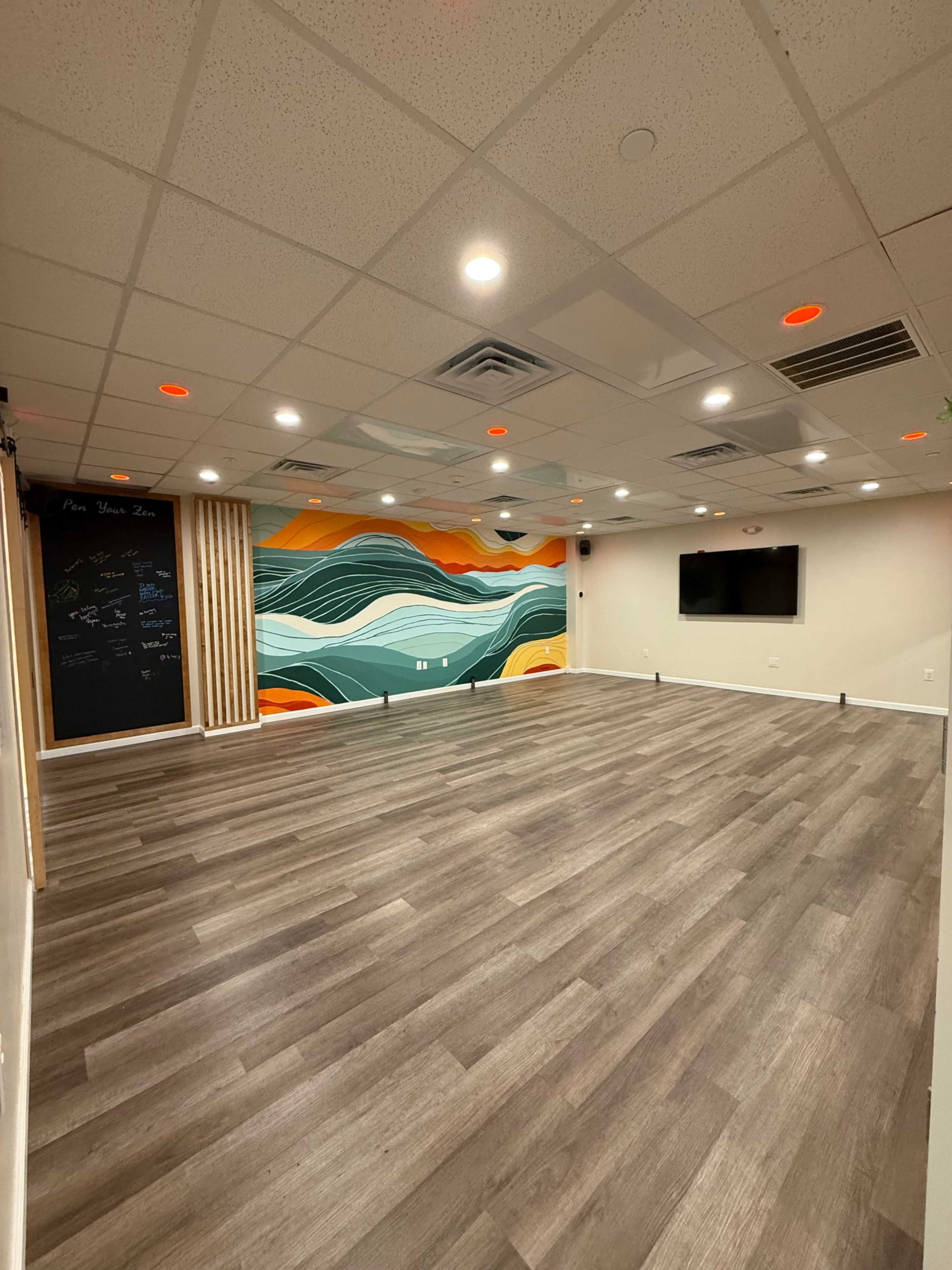The image shows a spacious, well-lit room with a wooden floor, featuring a colorful mural on one wall and a television mounted on the opposite wall.
