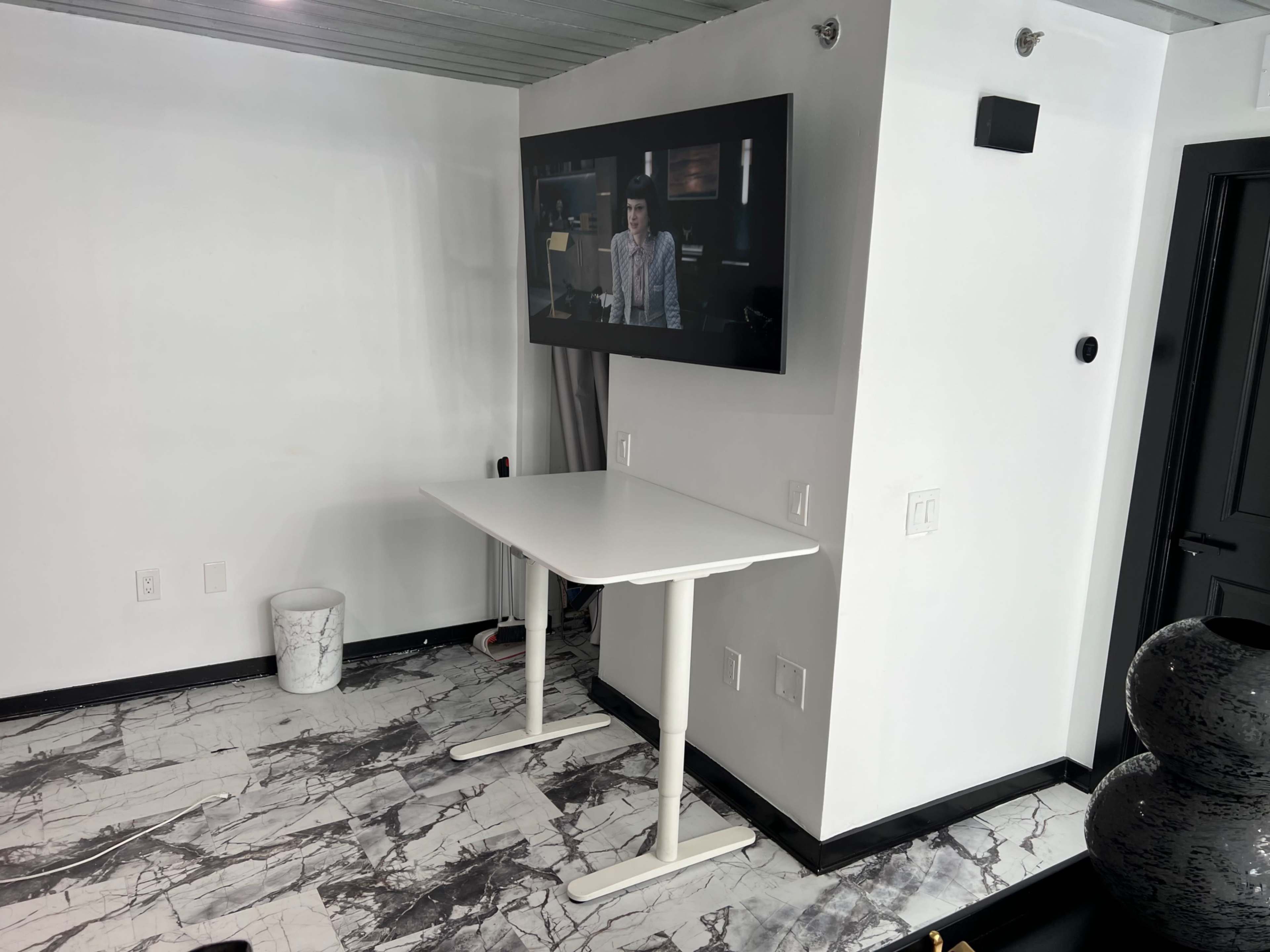The image shows a minimalist room with a white standing desk and a television mounted on the wall.