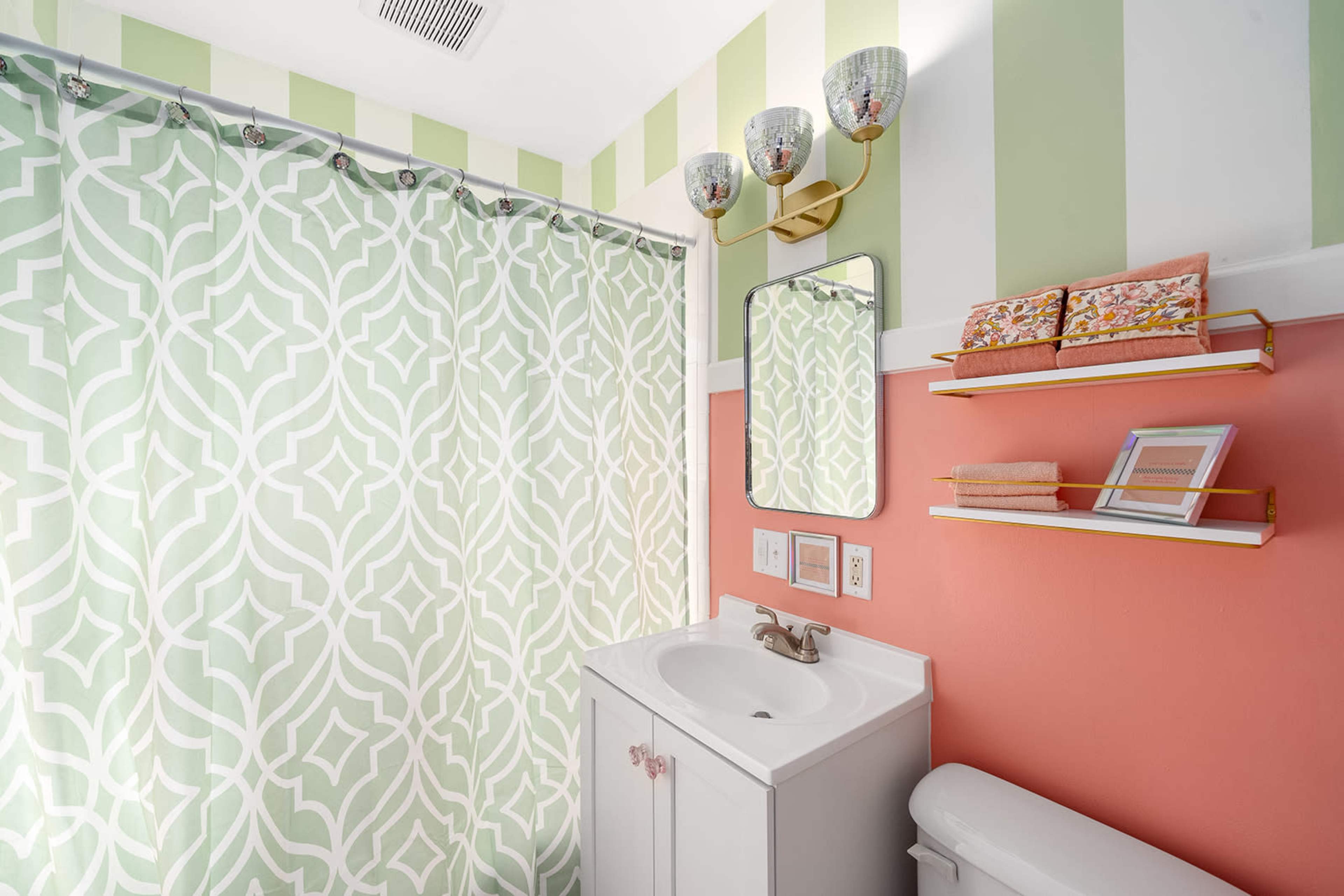 A bathroom with a patterned shower curtain, a white sink, a mirror, and floating shelves displaying decorative items against colored walls.