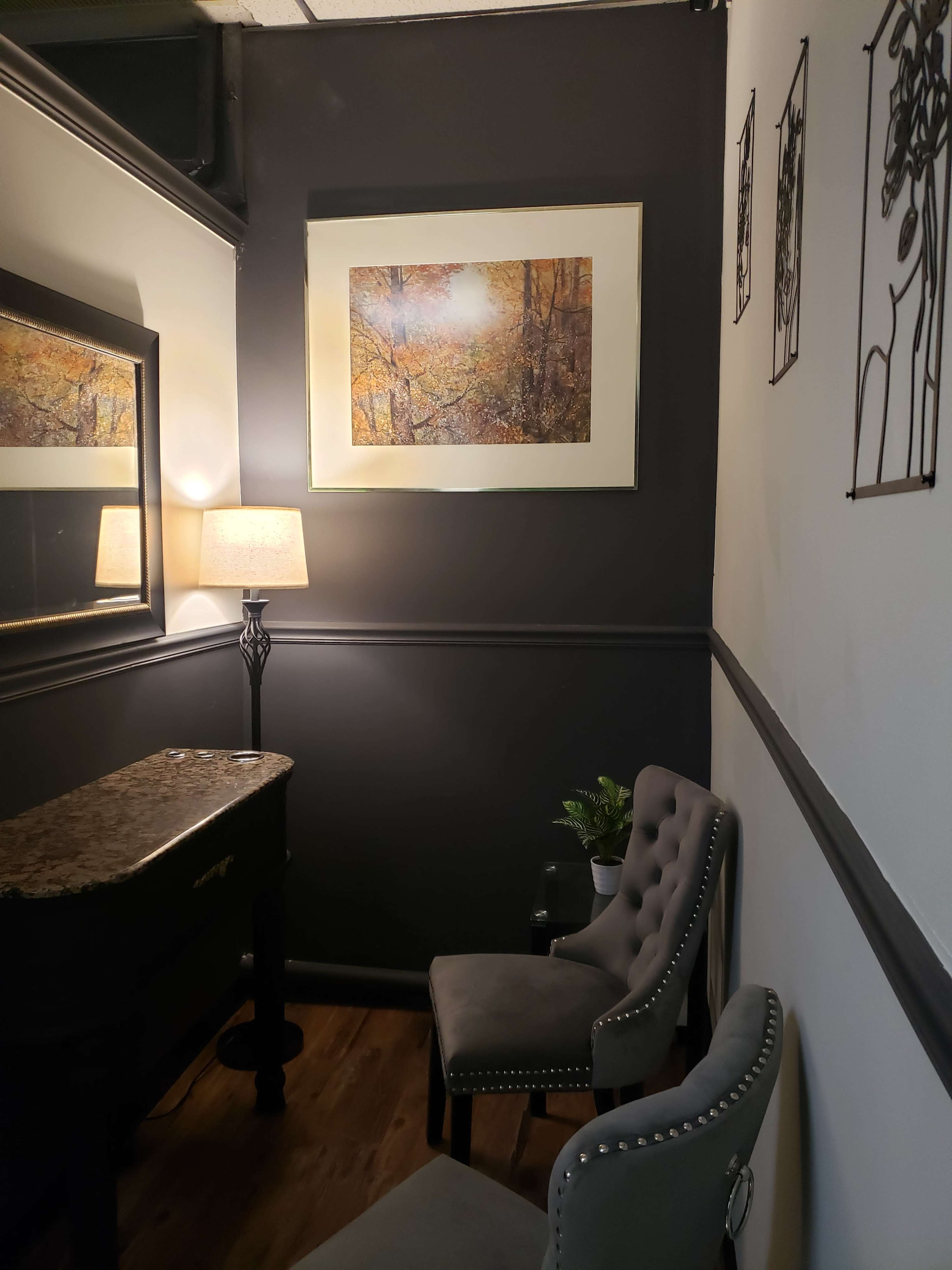 The image shows a corner of a room with two upholstered chairs, a lamp on a side table, a decorative mirror, and framed artwork on the walls.