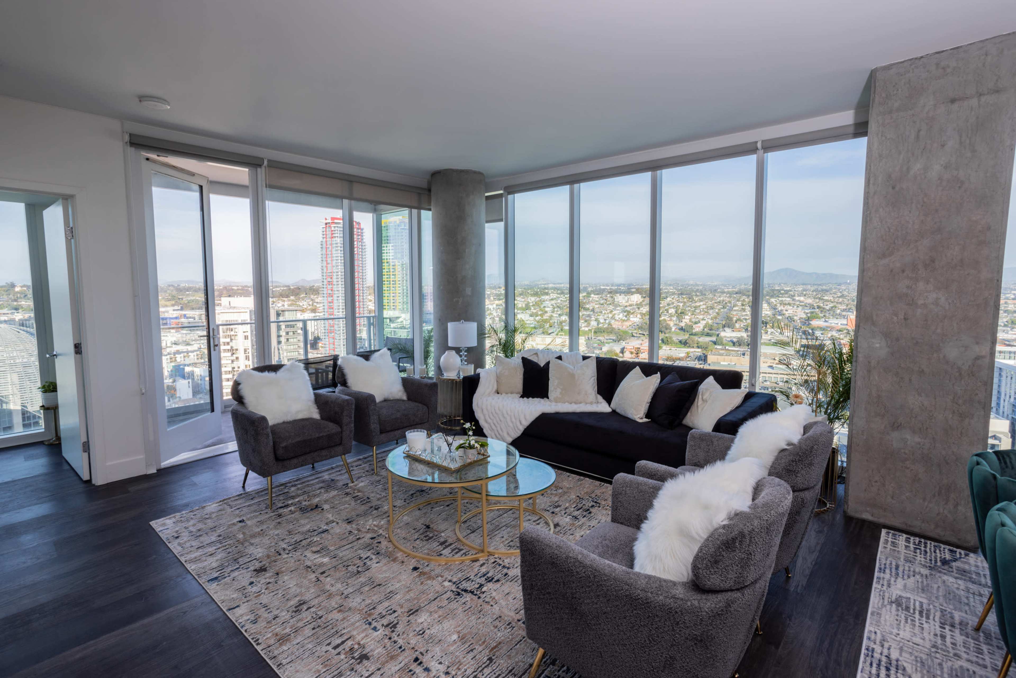 A modern living room with a large window offering a view of the city, featuring a black sofa, gray armchairs, and a glass coffee table.