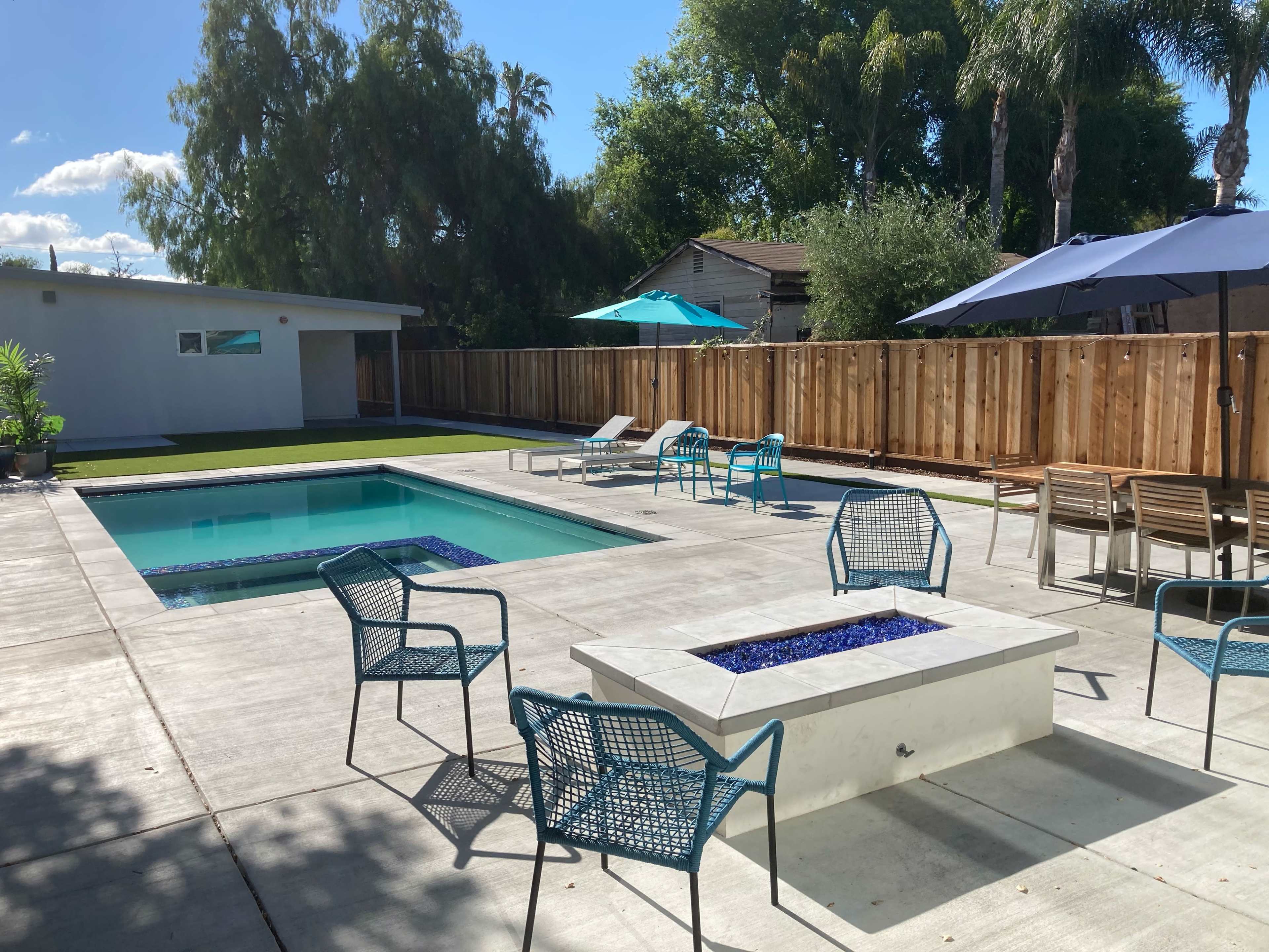 A backyard with a swimming pool, a fire pit, blue chairs, umbrellas, and a green lawn surrounded by a wooden fence.