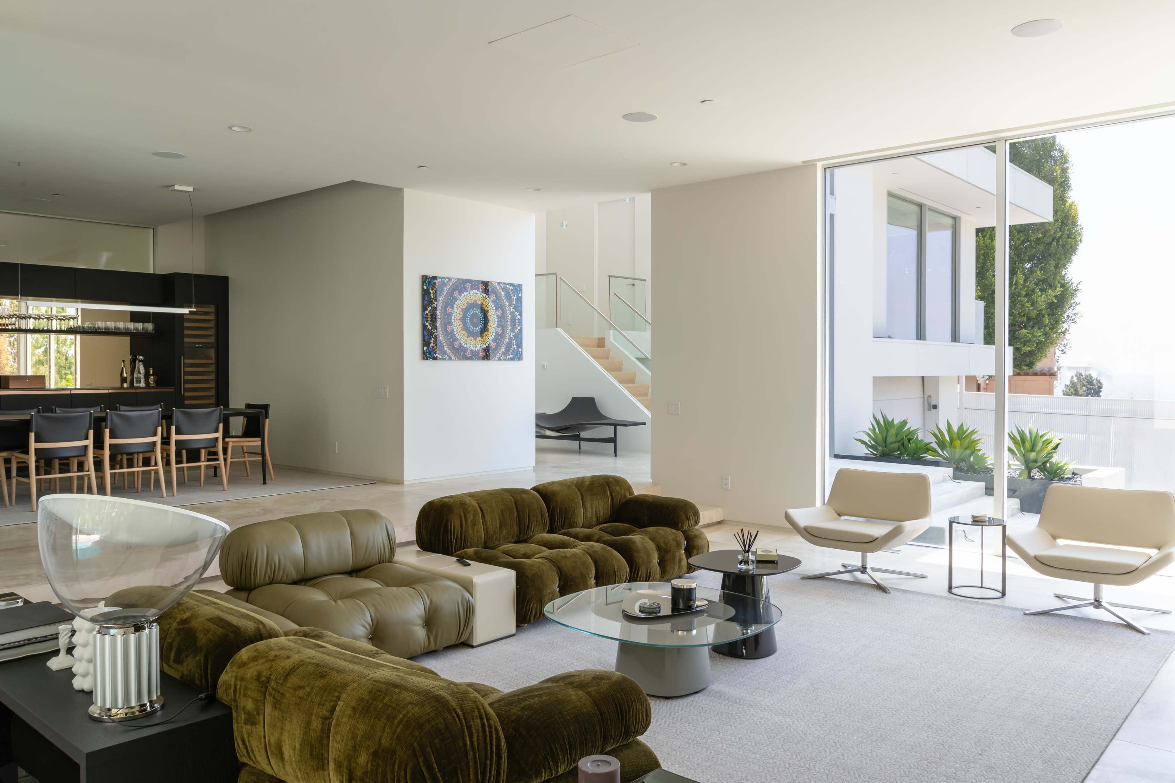 The image features a modern living room with a green velvet sectional sofa, a glass coffee table, and two white armchairs, alongside a dining area with a dark kitchen in the background.