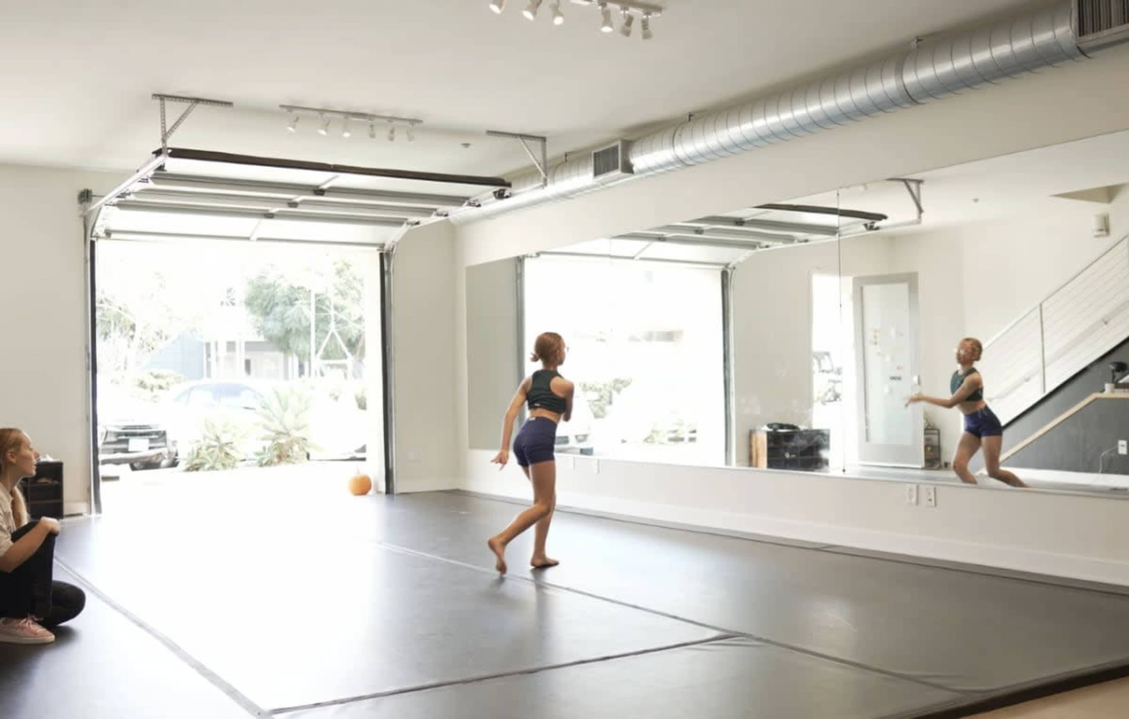 A dancer performs in a spacious studio with mirrored walls and large garage doors, while an observer sits on a mat.