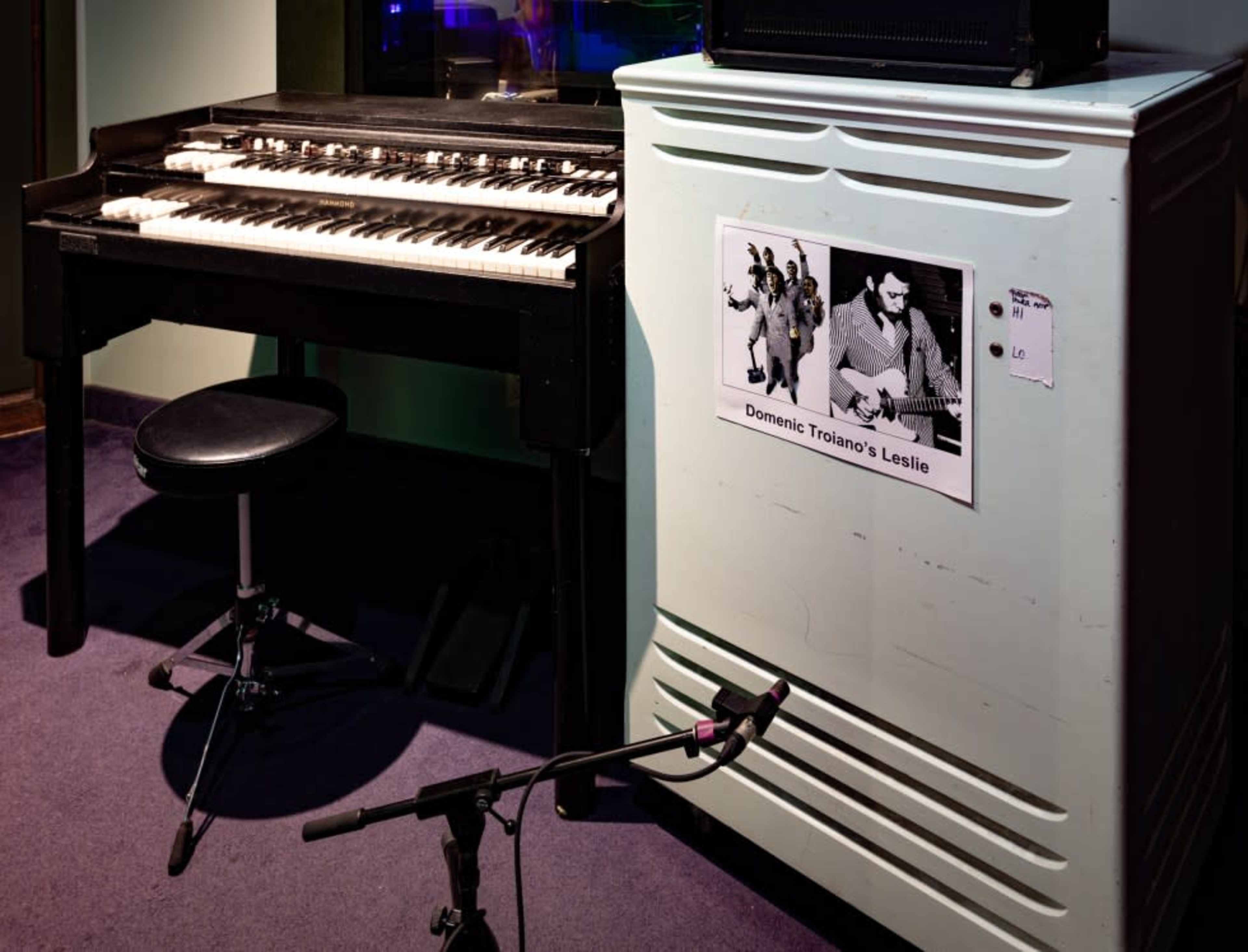 The image shows a vintage organ next to an amplifier, with a microphone positioned nearby and a poster of a musician attached to the amplifier.