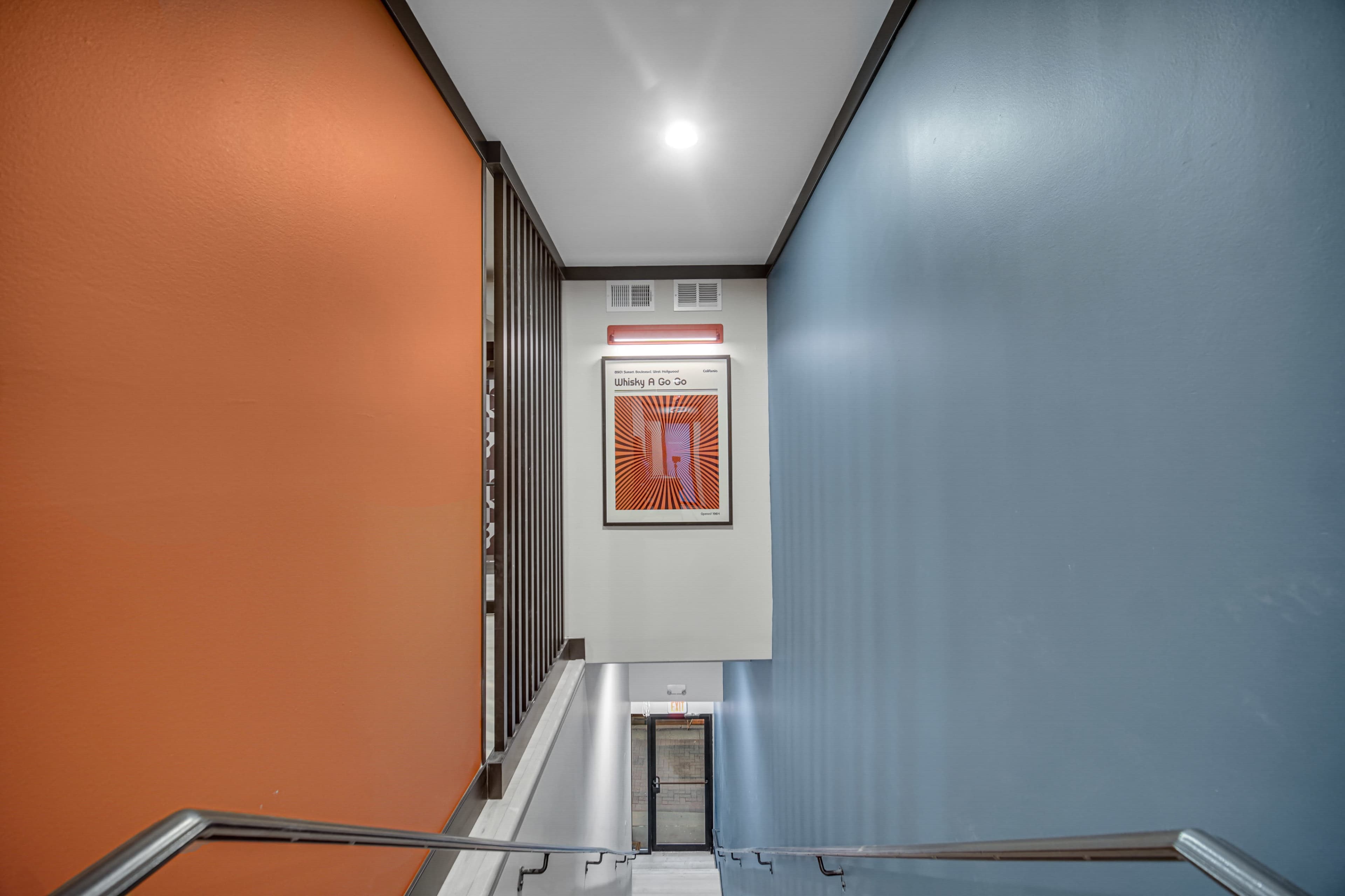 A staircase with orange and blue walls leads down to a door, featuring a framed artwork on the wall at the landing.