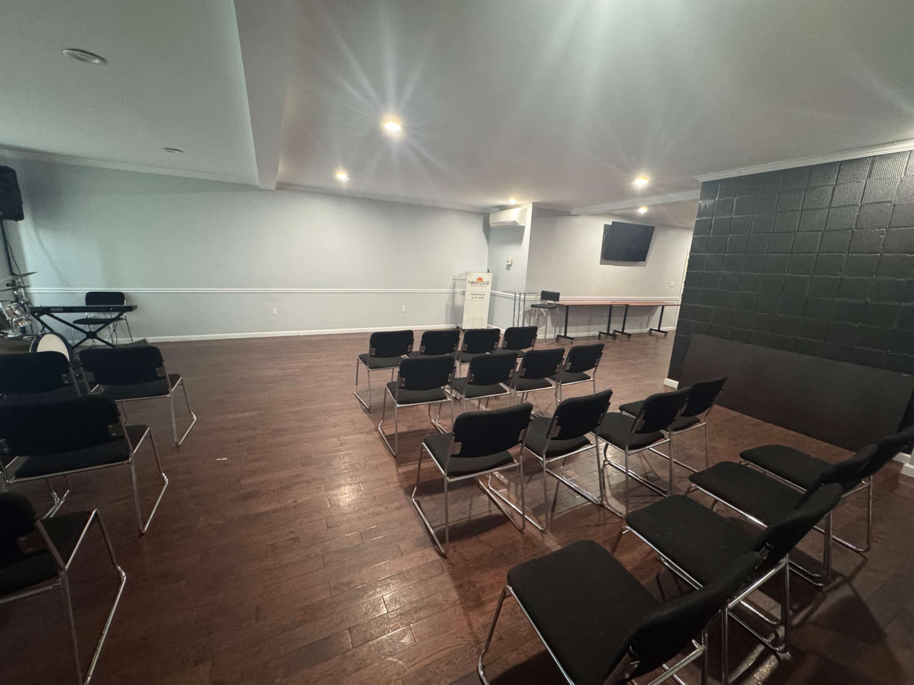 An empty room with wooden floors and rows of black chairs facing a blank wall, equipped with a small stage and a television.
