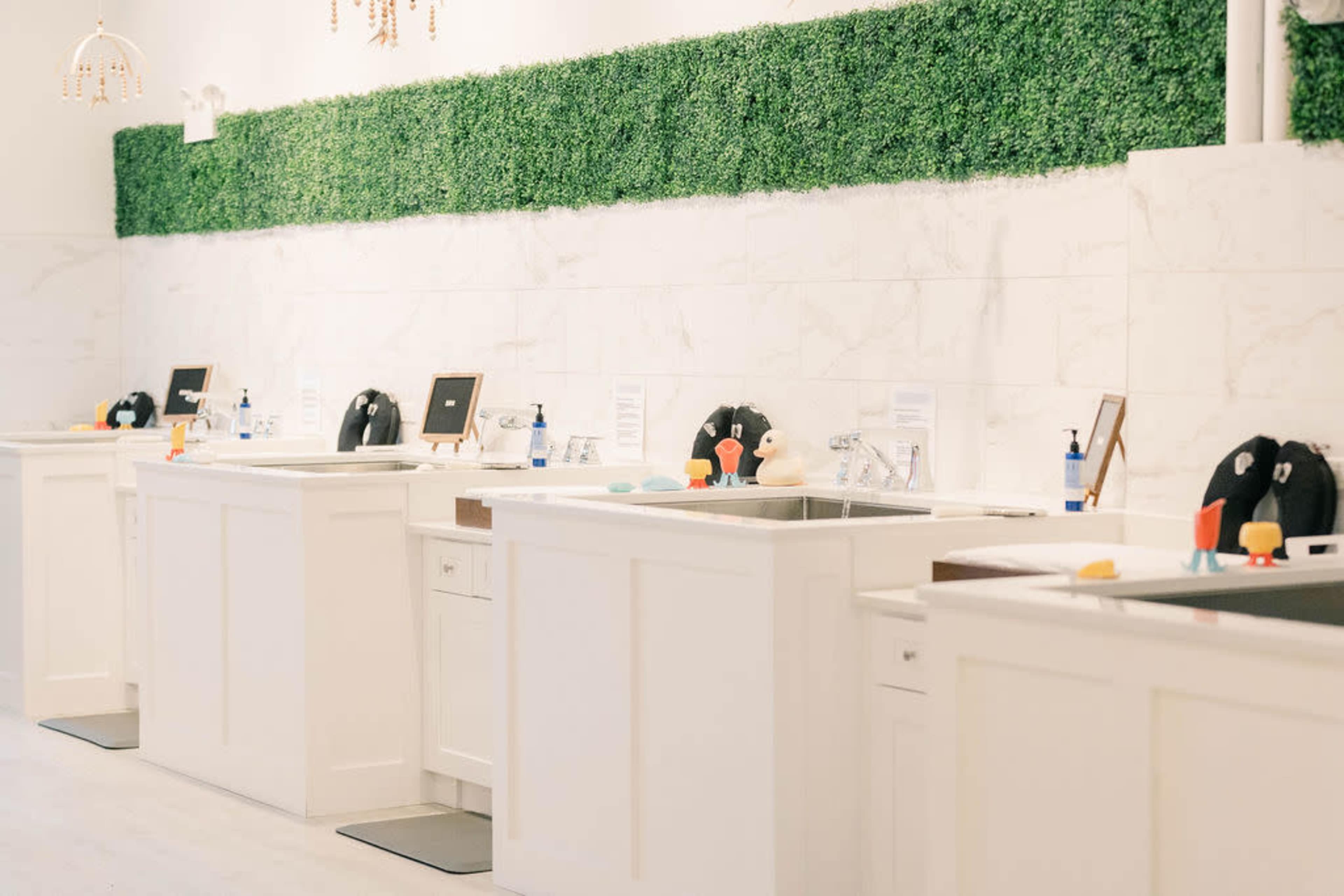 A clean, white-walled space with several white sink stations arranged in a row, each equipped with supplies and decorated with a green hedge backdrop.