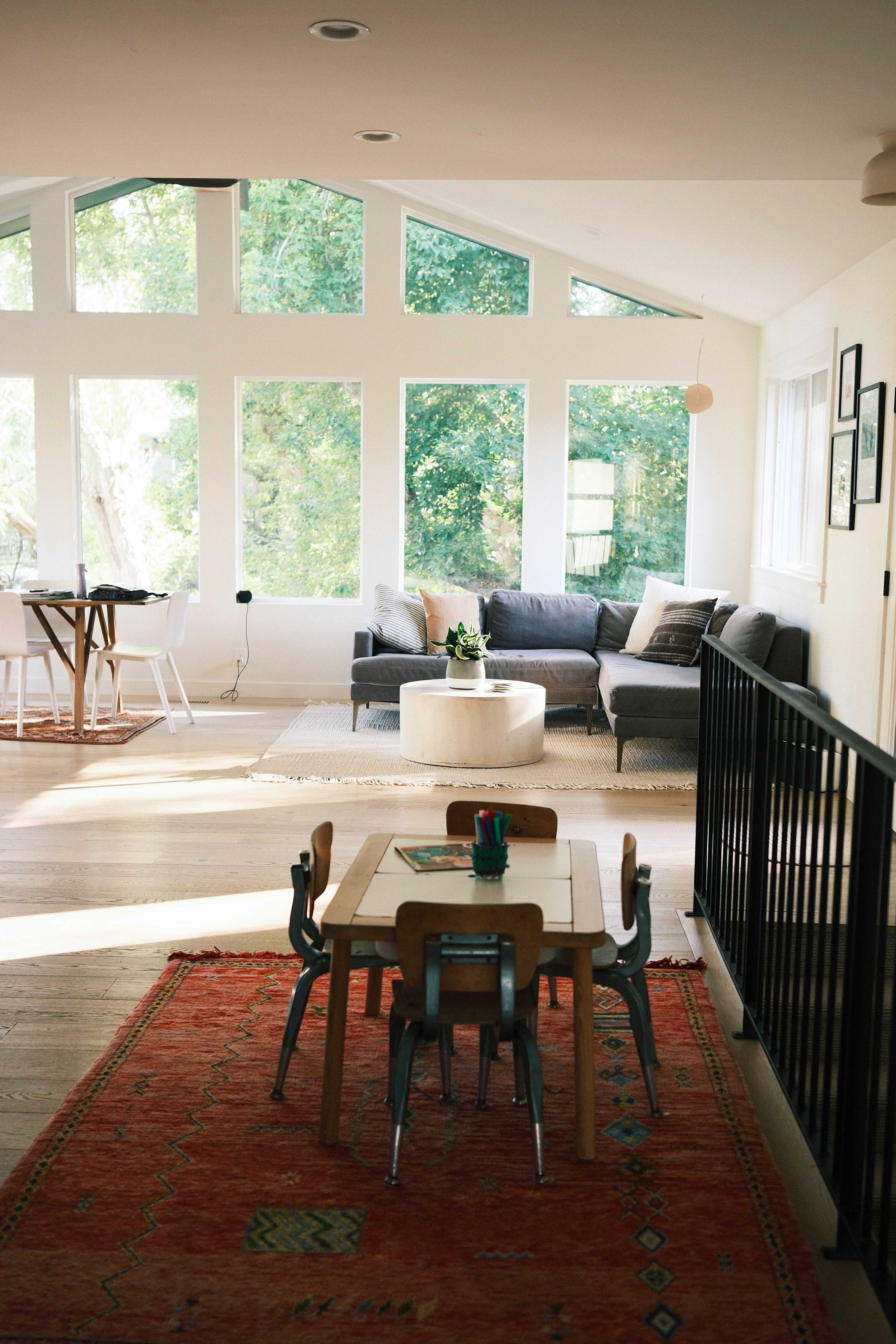 A modern open-concept living space features a small dining area with a table and chairs, a sofa in front of large windows, and a area rug on the wooden floor.