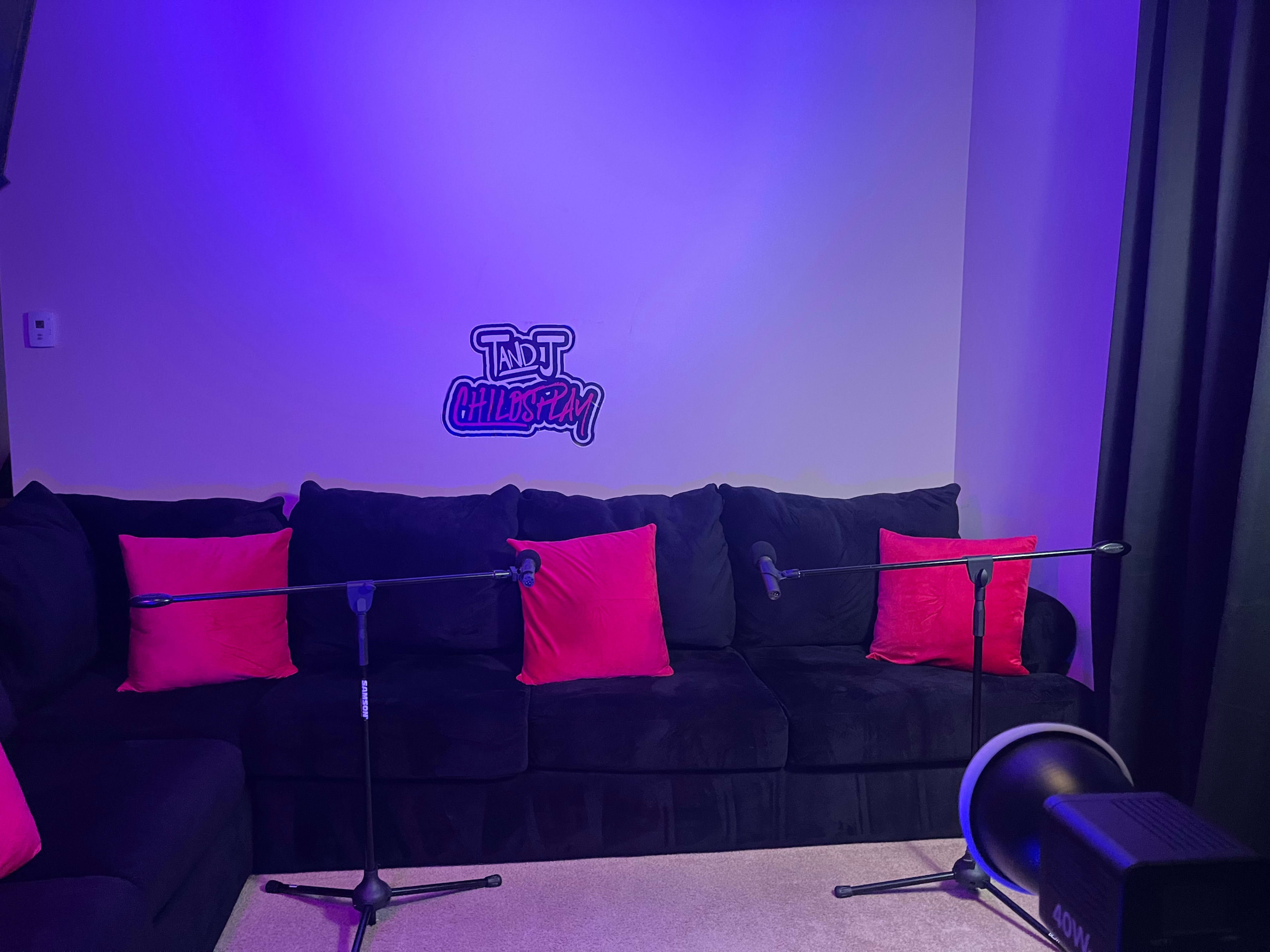A black couch with pink pillows is set against a wall featuring a sign that reads "TND ChillSpot," and two microphones are positioned on stands in front of it.
