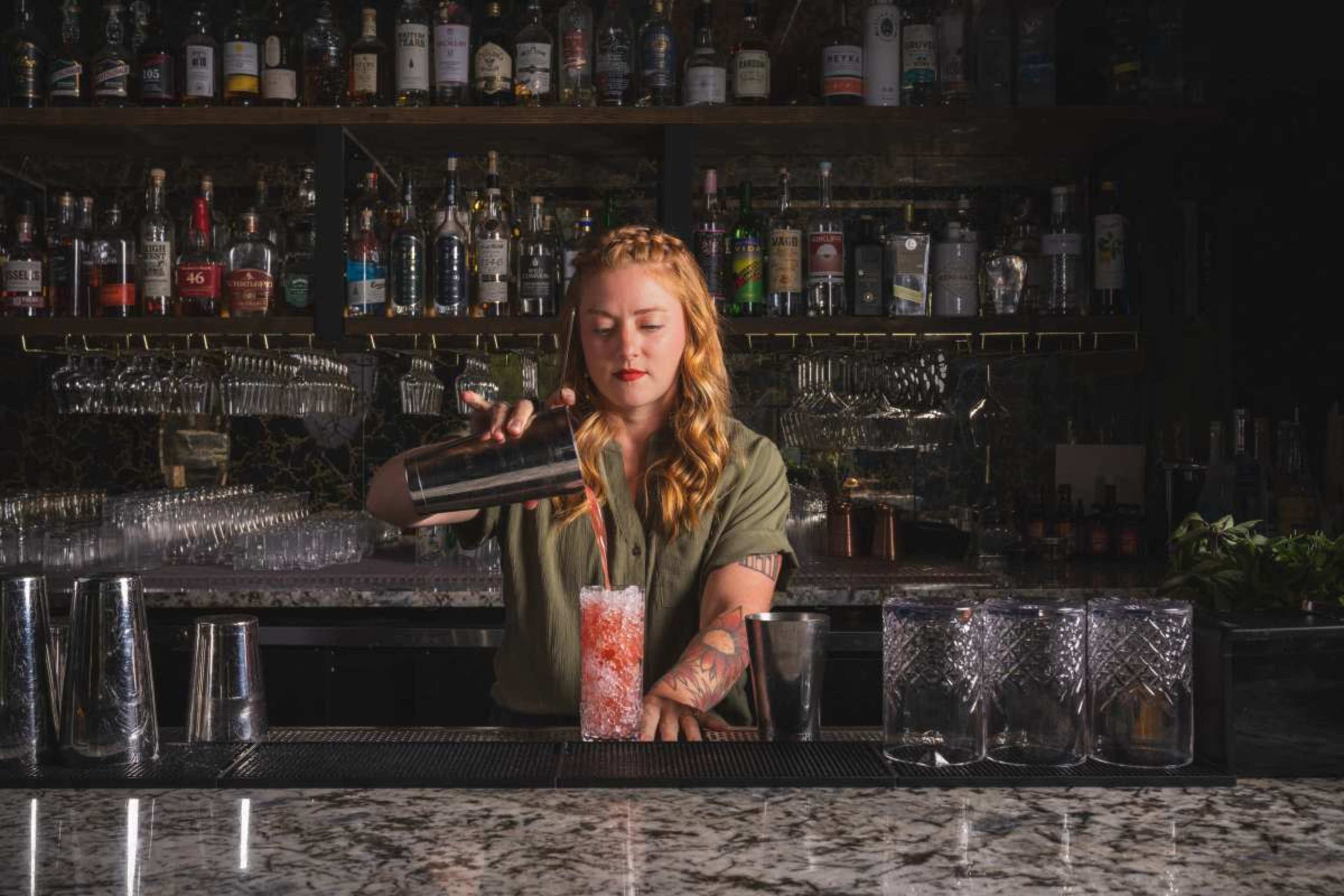 A bartender with braided hair is pouring a cocktail from a shaker into a glass filled with ice at a well-stocked bar.