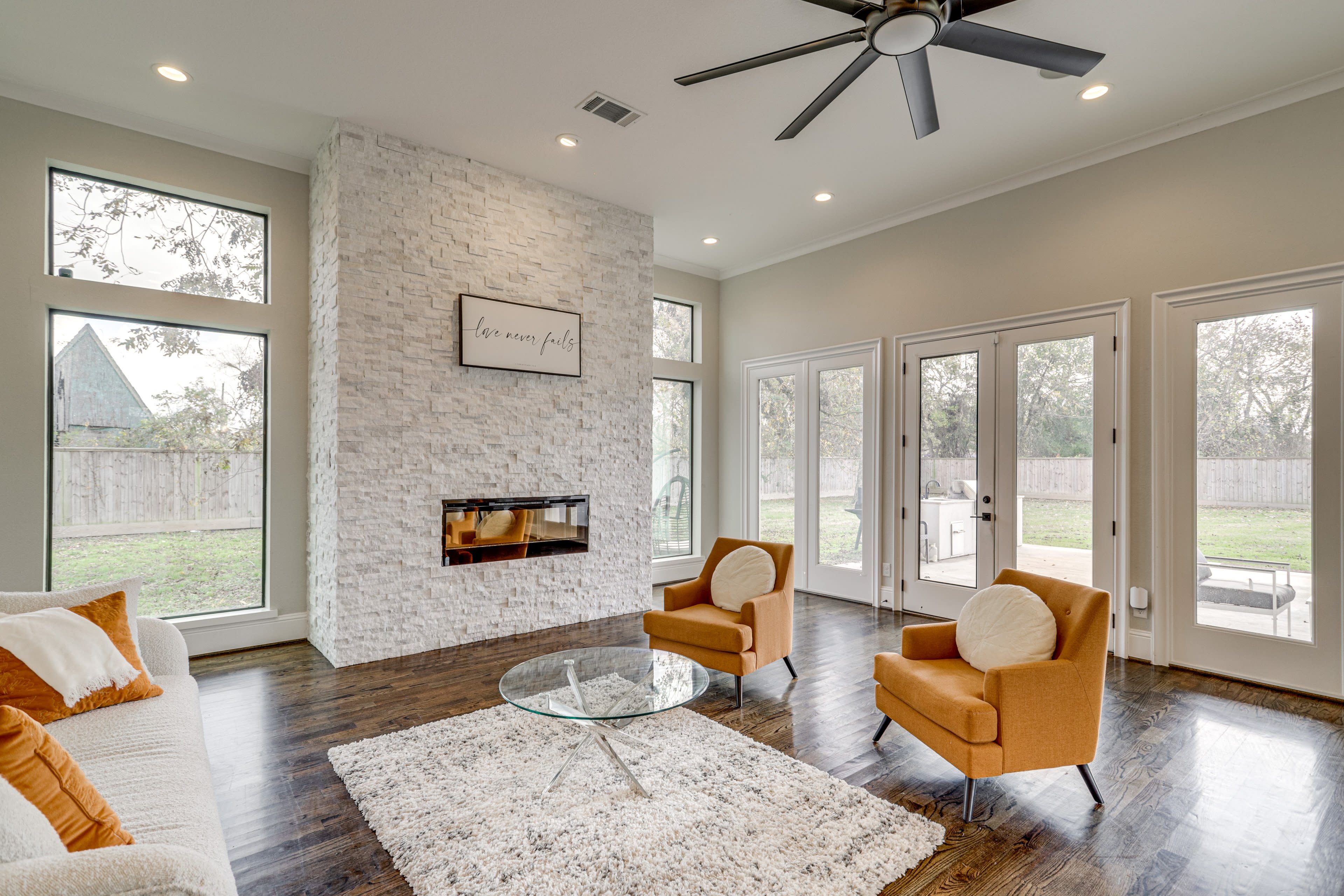 A modern living room features a stone fireplace, large windows, a round glass coffee table, and two orange armchairs.